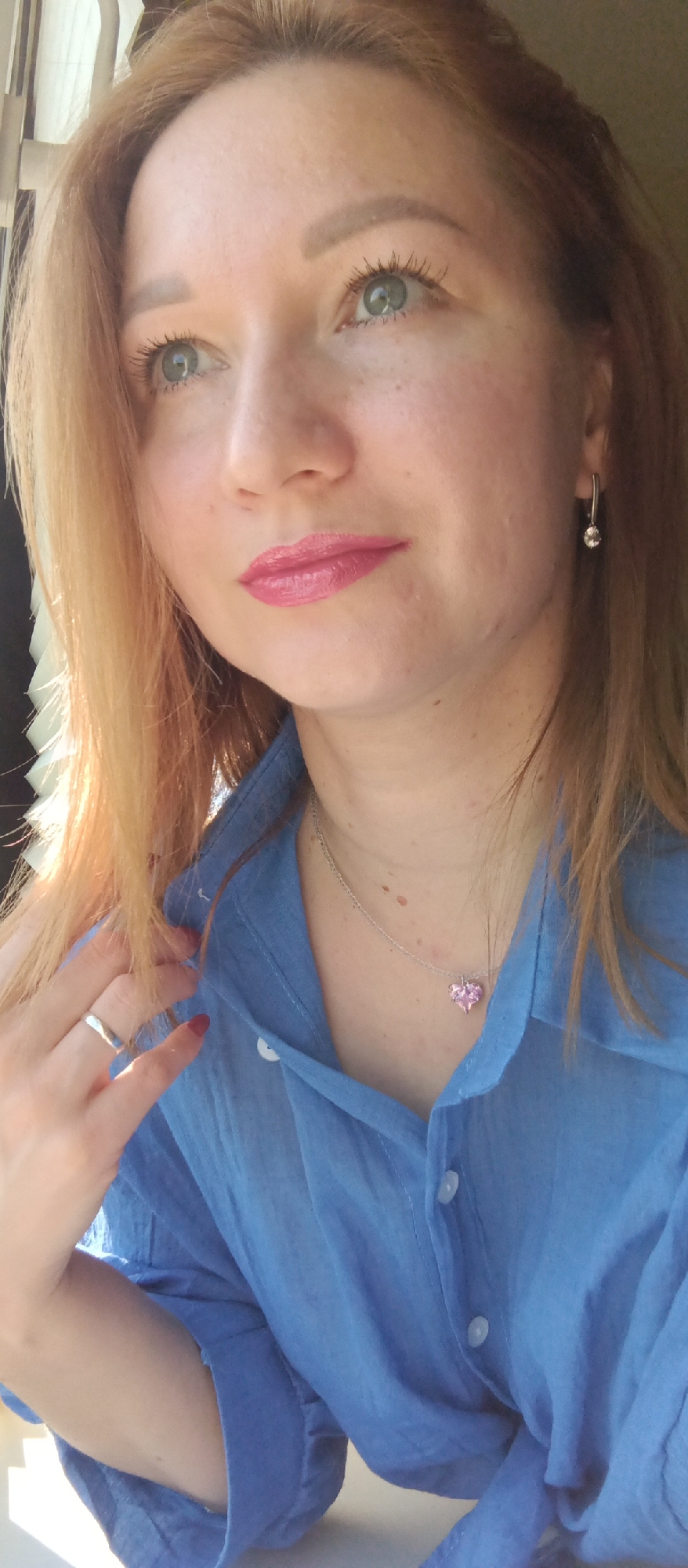 A medium shot depicts a fair-skinned woman with light red hair, her gaze directed slightly upward. She wears a blue button-down shirt, partially unbuttoned, revealing a silver necklace with a pink heart pendant. A silver ring adorns her left ring finger, and matching silver earrings dangle from her ears. Her lips are coated with pink lipstick. The background features vertical blinds, suggesting she is indoors, potentially near a window. No specific details indicate the city or location within "Unknown, Unknown."