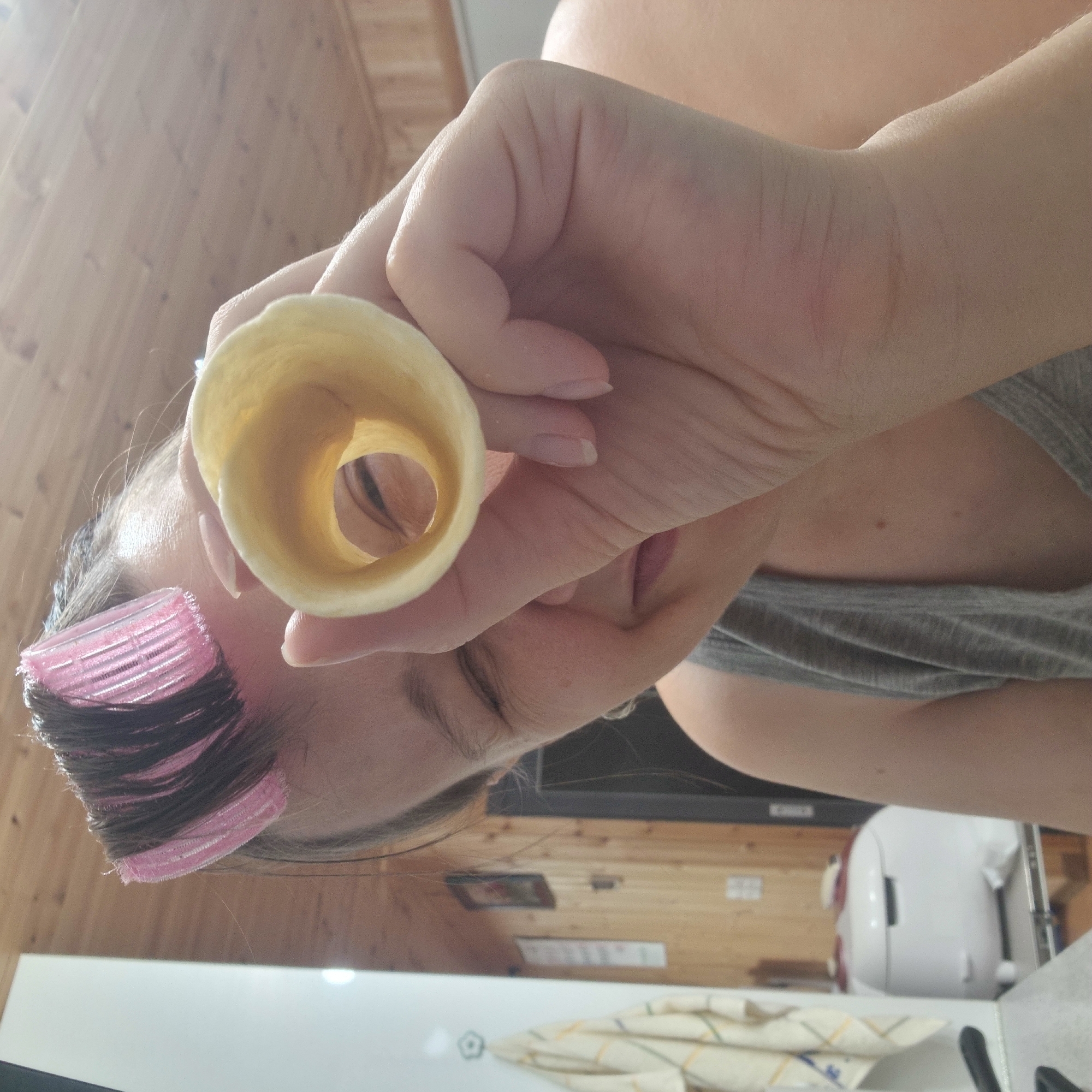 The image shows a close-up of a person's face, partially obscured by their hand holding a wafer cone up to their eye. The person has pink hair curlers in their hair and is wearing a gray tank top. The background appears to be a room with wood-paneled walls. On the left side of the image, a white appliance and a towel hanging on the wall are visible. The person's face is angled away from the camera, and they are looking through the cone. The lighting is bright, suggesting an indoor setting. There is no discernible information indicating the location in Dunnae-myeon, South Korea.