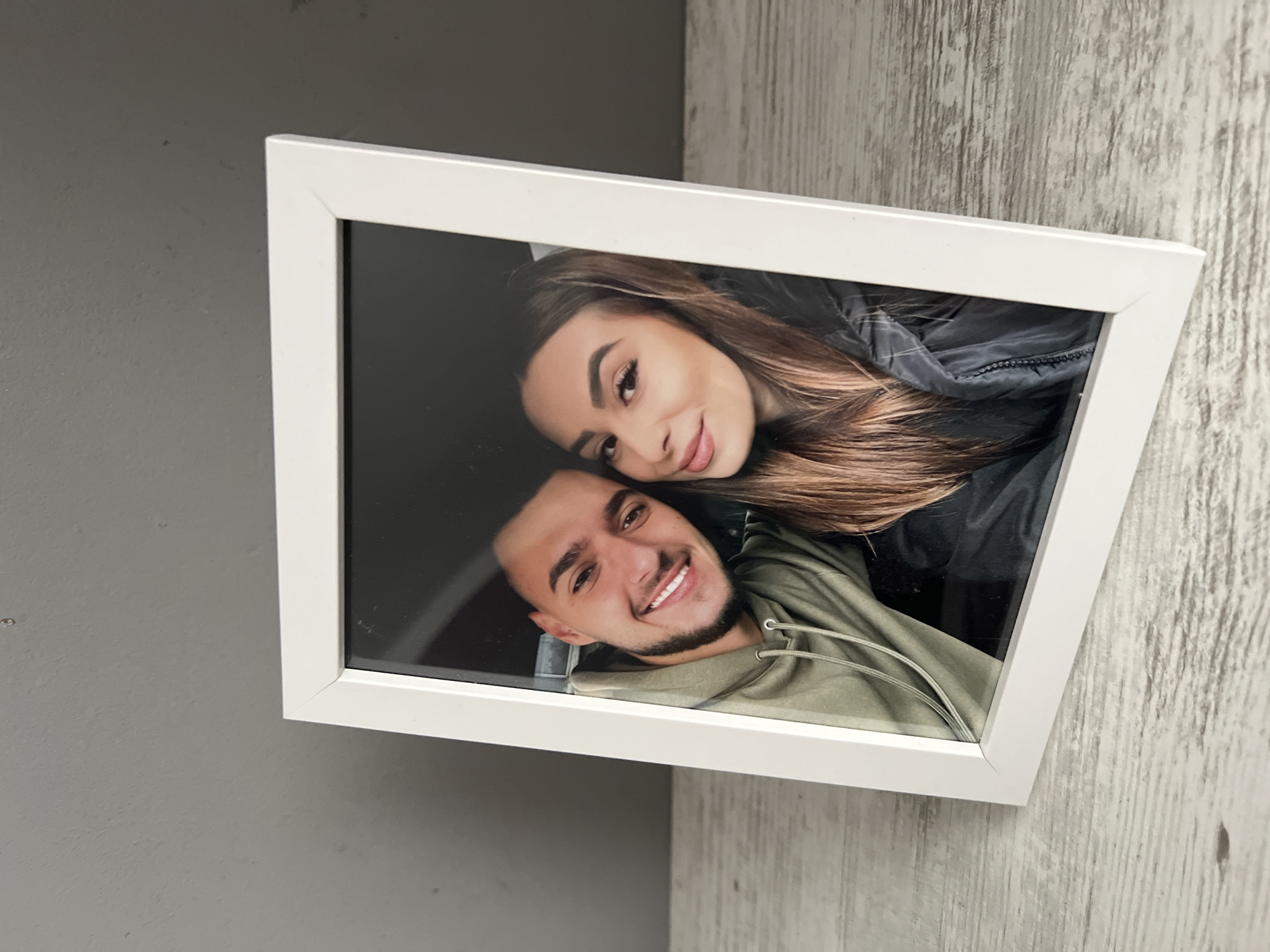 The image displays a white-framed photograph, captured at an oblique angle, featuring two young individuals, a man and a woman, smiling. The overall scene suggests the framed picture is hung indoors against a wall in Plovdiv, Bulgaria.
Within the photograph, a young woman is positioned on the right, her head slightly tilted. She has long, dark brown hair, styled to fall over her shoulder, and a bright, joyful smile. She is wearing a dark, possibly padded, jacket. Closely beside her, on the left, is a young man, also smiling broadly, revealing his teeth. He has short dark hair and a neatly trimmed beard. He is dressed in a green hooded sweatshirt with visible drawstrings. Their close proximity, with their cheeks almost touching, conveys a sense of affection and happiness.
The frame itself is a simple, white rectangle, angled as if mounted somewhat askew or photographed from an angle. The background behind the frame consists of two distinct wall sections. To the left, the wall is a solid dark gray or charcoal color, showing some minor blemishes. To the right, and extending behind the upper right portion of the frame, is a light-colored wall with a vertical wood-grain texture, suggesting a corner or a section where different wall treatments meet. There is no visible text in the image.