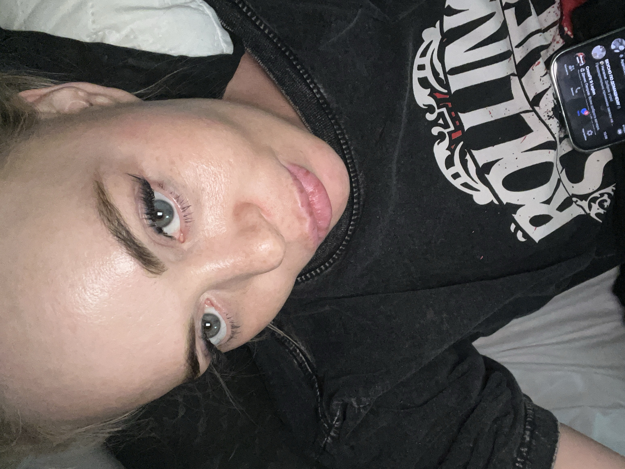 A woman with blonde hair is lying down and looking up at the camera. She is wearing a black t-shirt with a graphic of the Rolling Stones logo. Her makeup includes eyeliner and mascara. Her skin appears slightly oily. A smartphone is visible in the lower right corner of the image, showing notifications. The background is a bed with white and gray bedding. The scene is indoors.