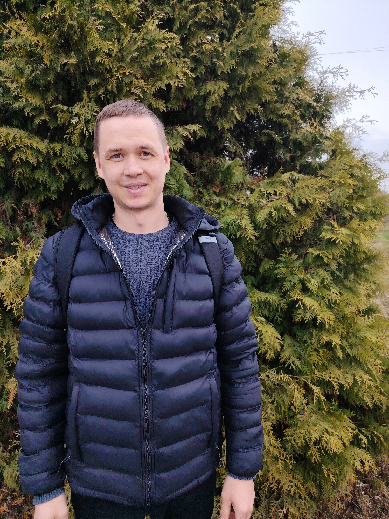 A man stands outdoors in front of a large, dense evergreen tree. He is wearing a dark blue puffer jacket over a textured gray sweater. The jacket is zipped up, and he has a black backpack slung over his shoulders. His hair is short and brown, and he is looking directly at the camera with a slight smile. The background is slightly blurred, suggesting the tree is close behind him. The lighting suggests an overcast day. The exact location is the city of Chaikovsky, Russia.