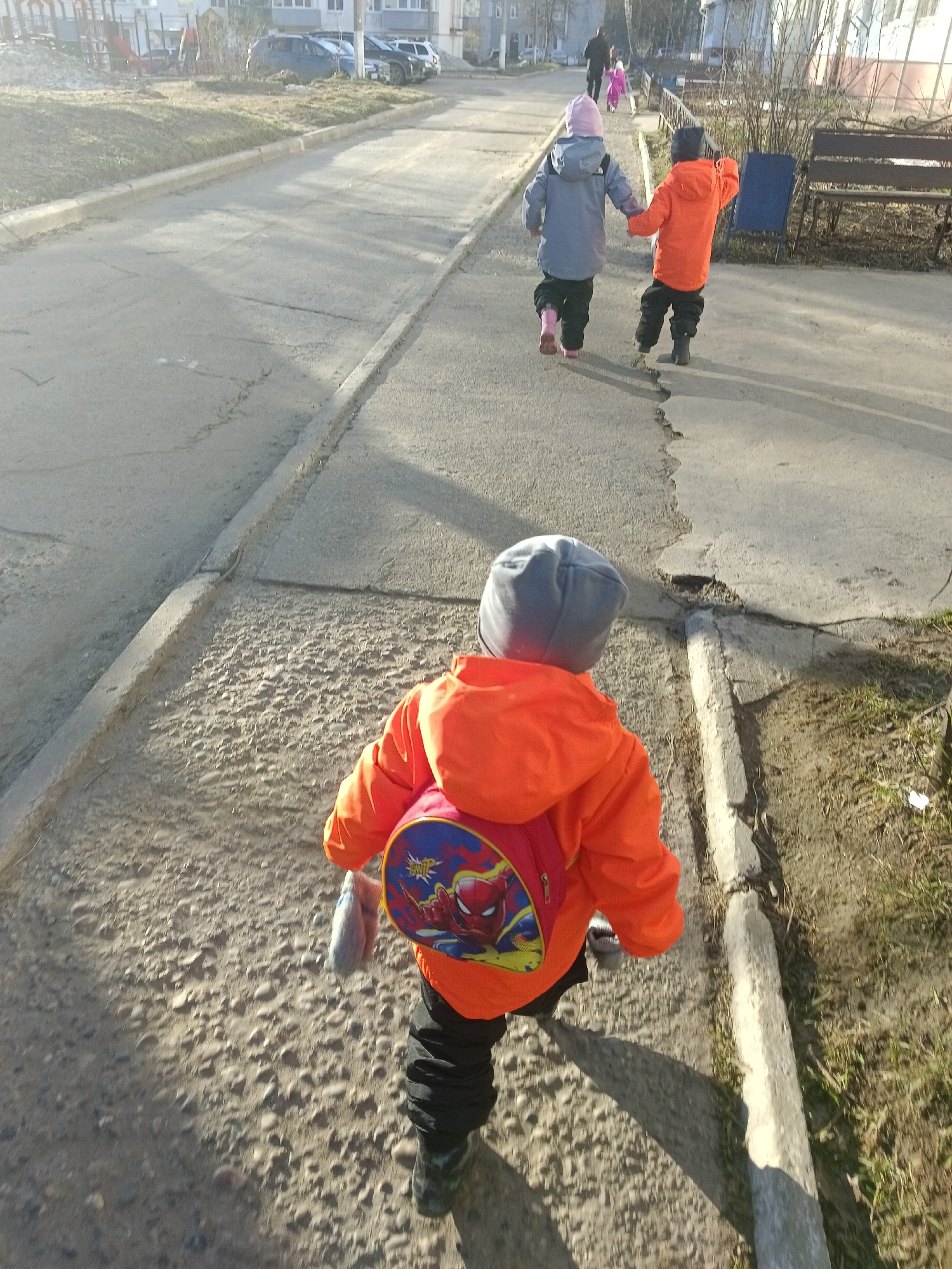 The photograph, taken from a high angle, features a group of children walking on a sidewalk alongside a paved road. A boy wearing an orange jacket and a gray hat with a backpack with a Spiderman print on it, walks toward the camera. Further ahead, two children, one in a gray jacket and pink hat and the other in an orange jacket, are walking away from the camera hand-in-hand. Benches are visible near the sidewalk. Cars are parked on the street in the background and apartment buildings. The photograph was taken in the city of Chaikovsky, Russia.