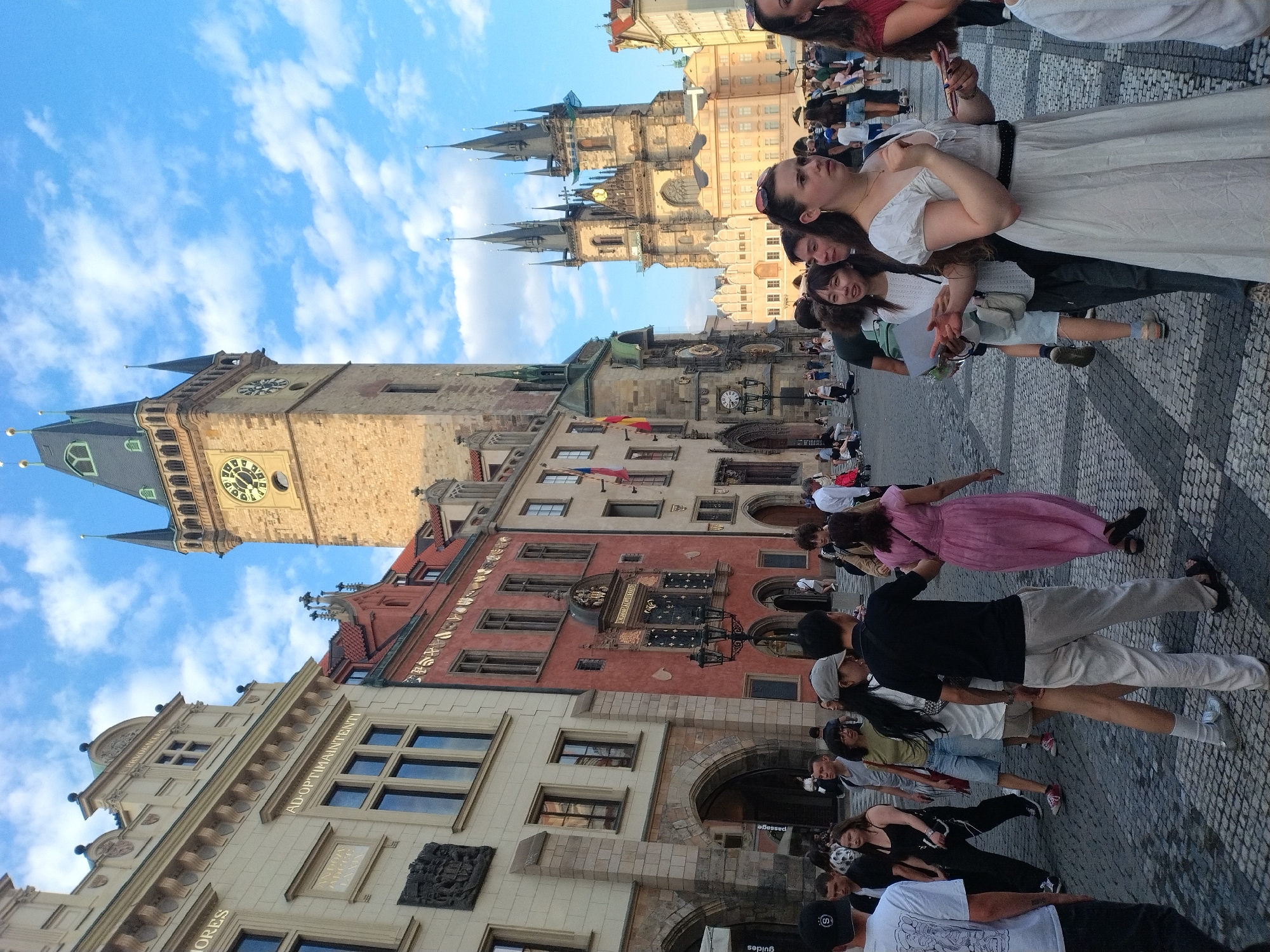 This is a medium-shot, eye-level, outdoor, daytime photograph taken in Prague, Czechia. The scene depicts the Old Town Square, with its iconic Astronomical Clock and the Church of Our Lady before Týn visible in the background. A large crowd of tourists and locals are milling about on the cobblestone square, some walking, some posing, and some gathered in small groups. The architecture is a mix of Gothic and Baroque styles, with ornate facades and clock towers. The sky is blue with scattered white clouds. The overall atmosphere is one of vibrant tourism and historical charm.