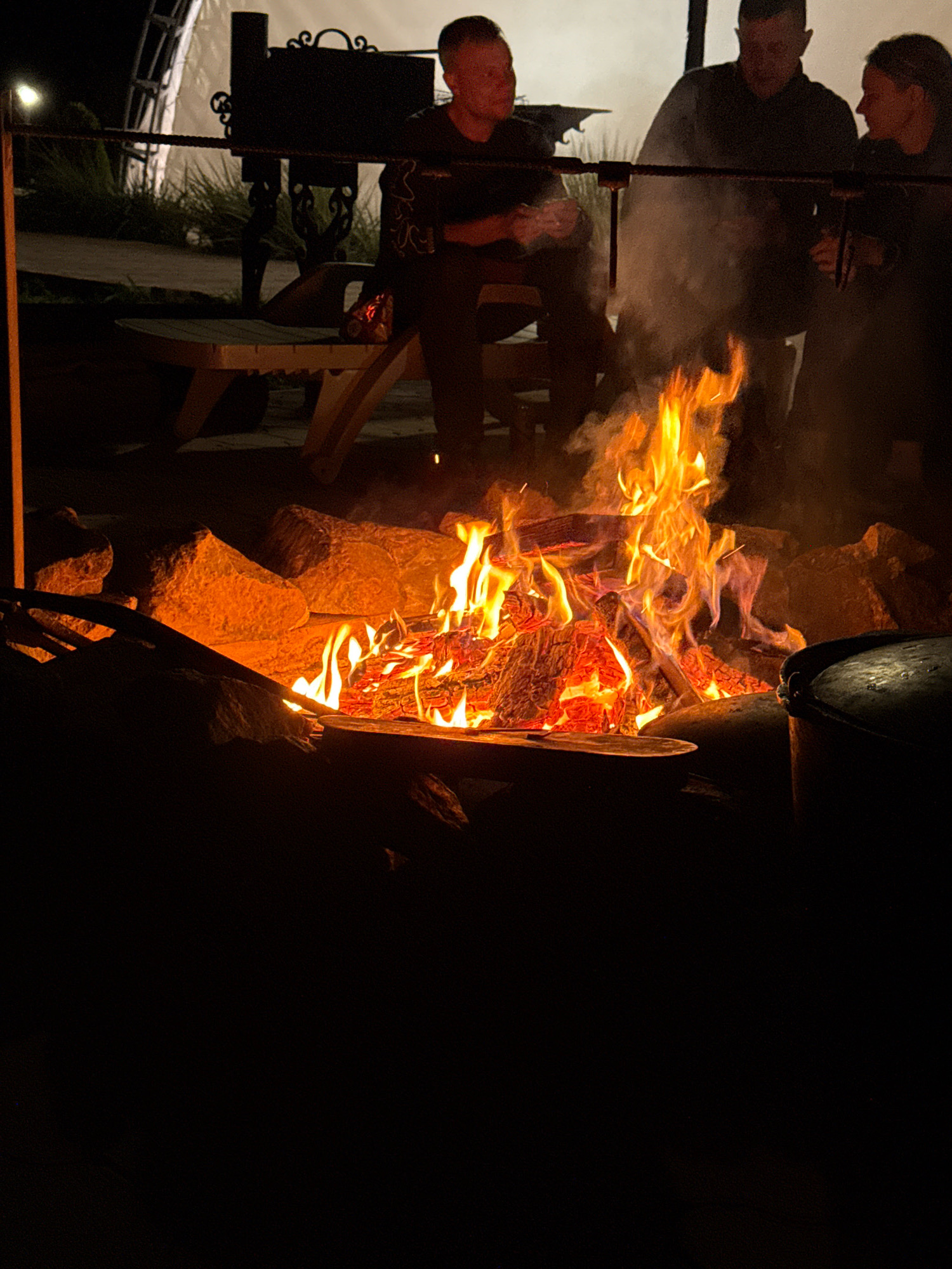In the city of Pakli, Russia, a group of three individuals are gathered around a bonfire at night. Two men and one woman are visible, their faces partially obscured by smoke and darkness. The man on the far left is looking down, holding something in his hands. The central man is looking towards the fire. The woman on the right is looking towards the fire as well, with her hands clasped in front of her. The bonfire is the primary light source, casting a warm glow on the surrounding rocks and logs. The environment appears to be an outdoor gathering area, possibly a park or campsite, with a picnic table and some vegetation visible in the background. A metal pot or cauldron is also present near the fire. The overall atmosphere is one of a relaxed, communal evening gathering.