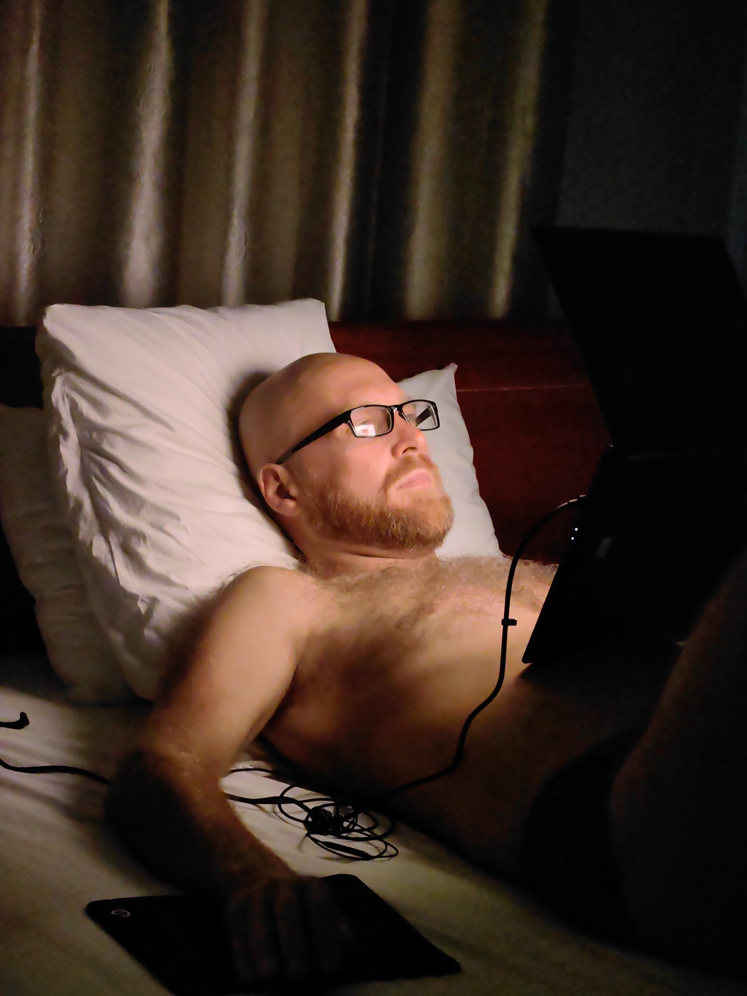A bare-chested, bald man with a red beard and glasses lies in a bed in Bai Chay Ward, Vietnam, propped against a white pillow in front of a wooden headboard, with curtains in the background. He appears to be looking up and to the right. He holds a dark electronic tablet in one hand and has a partially obscured laptop in front of him. Wires from electronic devices are visible on the bed.