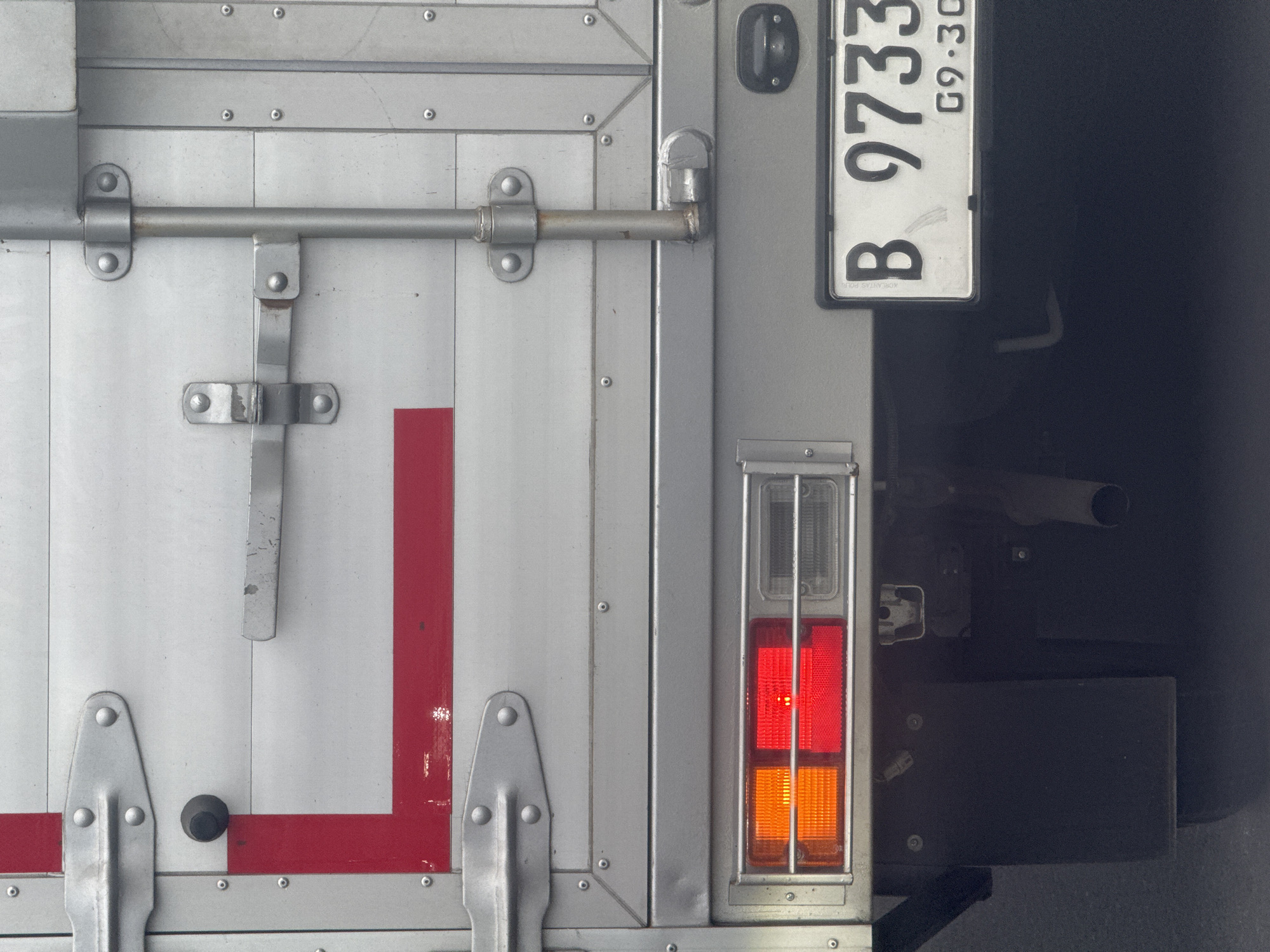 The image shows the rear of a truck in Nusa Dua, Indonesia. The truck is painted silver and has a red L-shaped stripe on the back door. The door has a metal latch and hinges. To the right of the door is a license plate with the number B 9733. Below the license plate is a tail light, with red and orange sections. The background is dark and mostly obscured.