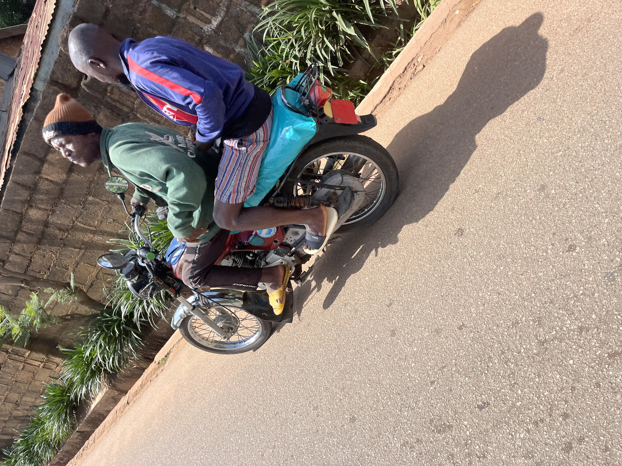 Two men are riding on a red and blue motorcycle on a paved road. The man in front is wearing a green sweatshirt and a brown and black beanie. The man behind him is wearing a blue jacket with red stripes. Both men are looking up and to the right, away from the camera. The motorcycle has a blue plastic cover on the seat. The road is lined with green plants and a brick wall. The scene appears to be in a city, possibly Gura Top, Nigeria.
