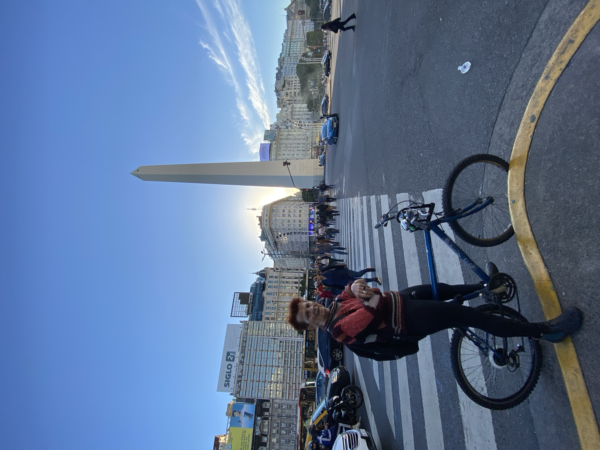 This urban landscape image, taken in Buenos Aires, Argentina, captures a lively street scene featuring the iconic Obelisco landmark under a clear blue sky with sparse, wispy clouds. The photograph appears to be taken during the late afternoon or early morning, evidenced by the warm, low sun casting a glow behind the Obelisco and creating subtle backlighting.
In the foreground, a woman with reddish-brown hair, dressed in a patterned red and black sweater and dark attire, smiles directly at the camera. She stands beside a blue mountain bike, which rests on a pedestrian crosswalk marked with white stripes. The asphalt road also features a prominent curved yellow line. A small piece of white litter is visible on the road surface near the bicycle.
The background showcases the bustling city, with the towering Obelisco positioned prominently on the left. Multi-story buildings, characteristic of a city center, line the streets. Numerous pedestrians are visible, some walking along sidewalks, others crossing the street on crosswalks. Various vehicles, including cars (a notable blue one), a bus, and motorcycles, populate the street. Traffic lights and streetlights are also present.
Visible text fragments on billboards and signs include "SIGLO 21 en" and "Lidere," suggesting a brand or institution. Another advertisement displays parts of phrases like "Te soy franc," "credito es," and "ratis posto" (likely "gratis"). The image conveys a dynamic urban environment with a personal touch from the smiling subject.