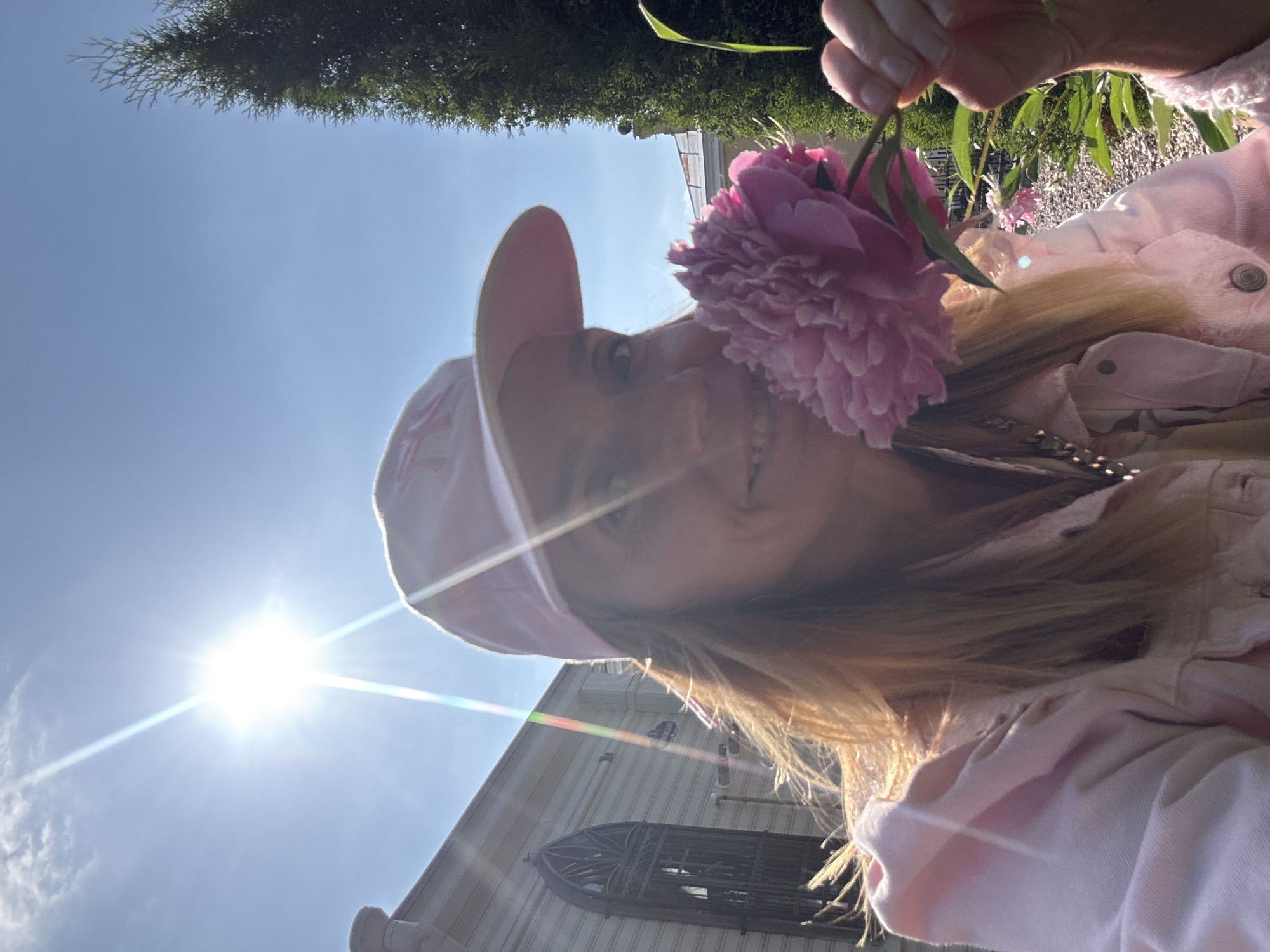 This media file is a portrait of a young woman with blonde hair, wearing a pink cap and a pink jacket. She is holding a pink peony flower up to the camera, obscuring part of her face. She appears to be smiling. The background shows a bright blue sky with a strong sun casting lens flare across the image. There is also a glimpse of a building with a decorative window and some green foliage, possibly from trees and bushes, suggesting an outdoor setting. The overall lighting indicates it is daytime, likely sunny and warm. The woman's pose and the flower suggest a playful or cheerful mood. The image appears to be a selfie.