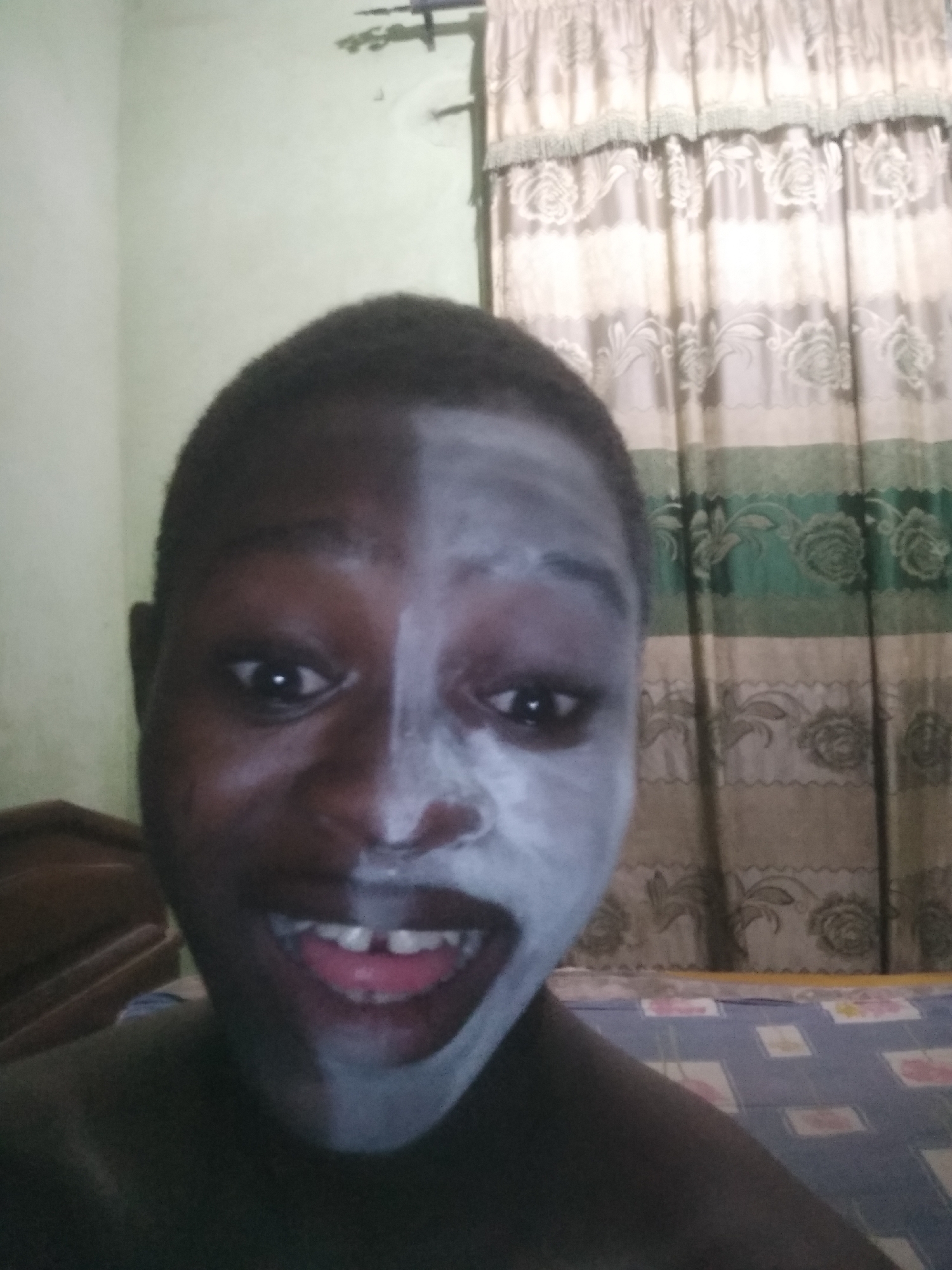 A close-up photograph features a dark-skinned person with half of their face covered in a white substance, possibly a cosmetic mask or cream. The person is smiling widely, revealing their teeth. The background includes a green and white patterned curtain and a bed covered with a blue patterned bedsheet. The location, Shangisha, Nigeria, cannot be confirmed from the image alone. The environment suggests an indoor setting.