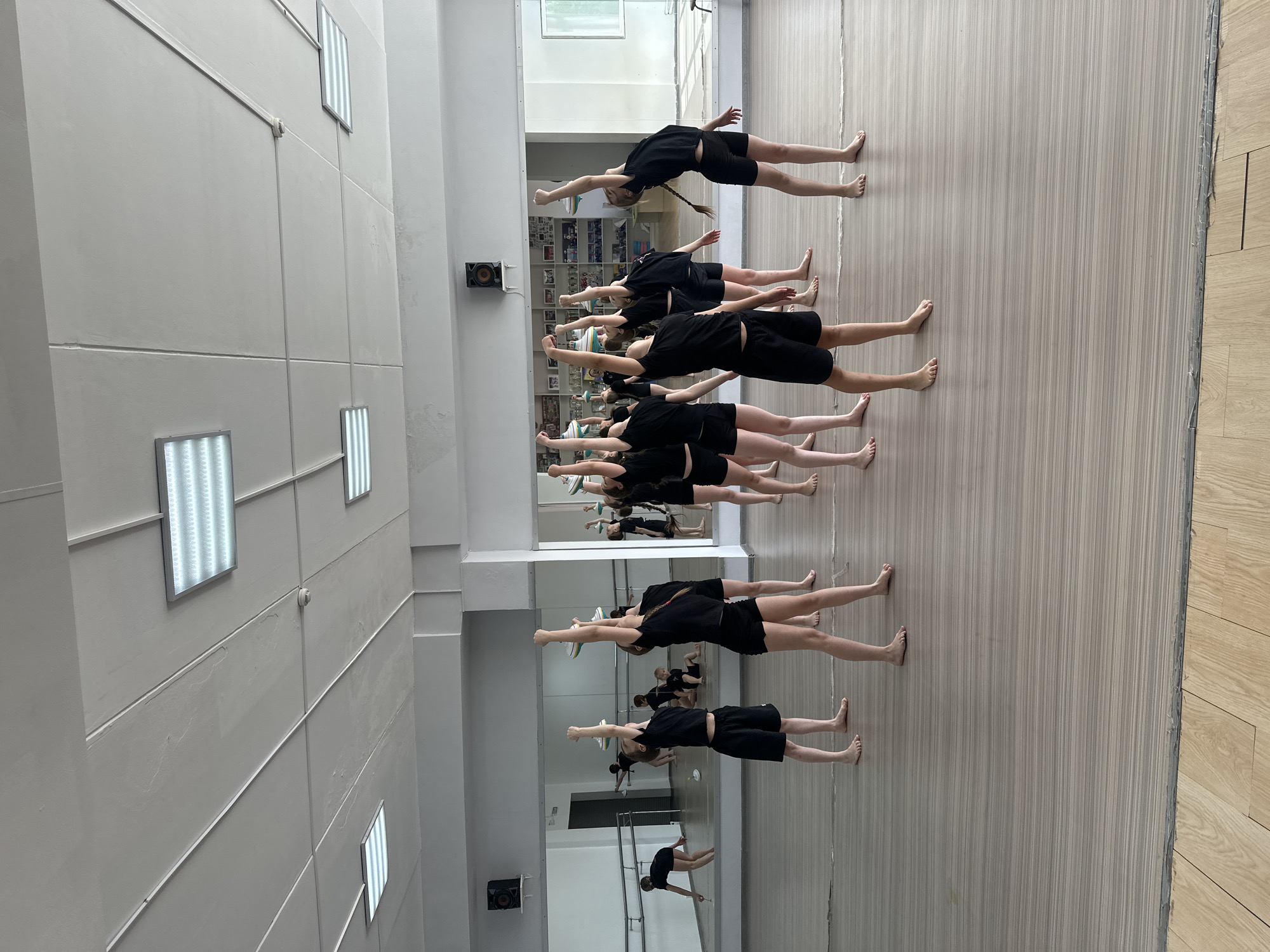 The image shows a group of approximately 10 young people, likely girls, in a large room with a wooden floor, possibly a dance studio or gymnasium, in Votkinsk, Russia. The individuals are dressed in black shorts and tops, and they are barefoot. They are positioned in a row, lying on their backs, with their arms raised overhead. A large mirror runs along one wall, reflecting the group and the surrounding space. The room appears to be well-lit, with rectangular fluorescent lights mounted on the walls and ceiling. The walls are white or light-colored. The angle of the photograph suggests it was taken from an elevated vantage point.