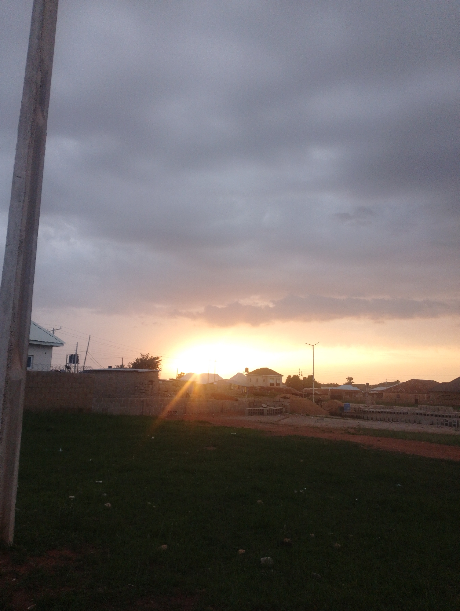 The image depicts a rural or semi-rural landscape in Katon Rikkos, Nigeria, during sunset. The sky is overcast with clouds, but the setting sun casts a warm, golden glow, creating a dramatic lighting effect across the horizon. In the foreground, a patch of dark green grass is visible, with some scattered debris. To the left, a tall, gray concrete pole stands prominently. Beyond the grassy area, a low brick wall forms a boundary.
In the middle ground, several buildings are scattered, some appearing to be under construction. These include houses with pitched roofs and a building with a light green roof. A street lamp with a single light fixture is positioned on the right side of the middle ground. The ground in this area appears to be a mix of dirt and construction materials, with piles of sand and stacks of bricks. Power lines and utility poles are also visible, suggesting some level of infrastructure. In the background, more houses and structures are discernible against the fading light. The overall scene conveys a sense of a developing community at the end of the day, with the natural beauty of the sunset contrasting with the ongoing construction. No people are clearly visible in the image.