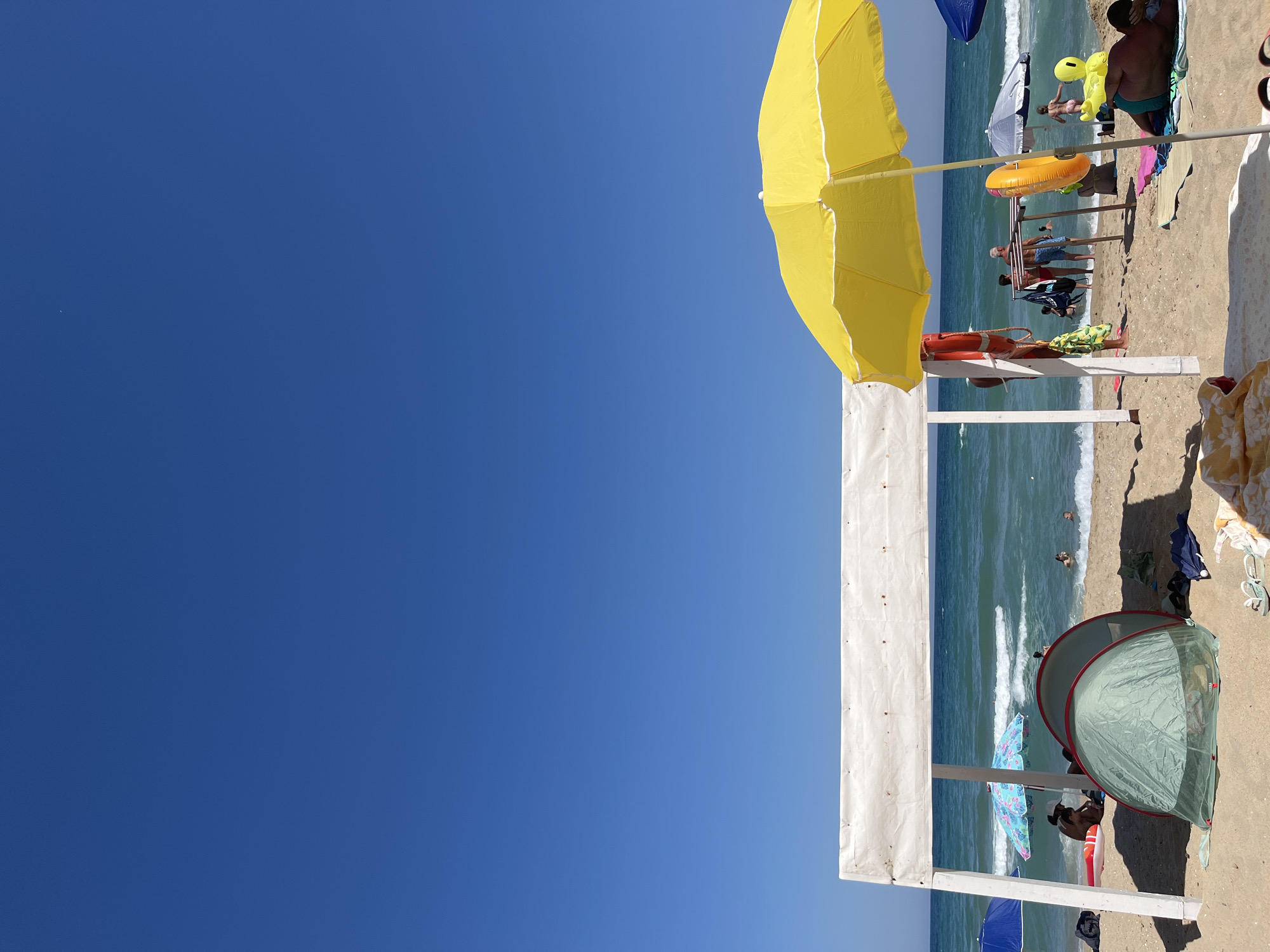 The image depicts a sunny day at a beach in Obzor, Bulgaria. The foreground is dominated by sand, with several beach accessories visible. A large, white, open-sided structure with a canvas roof stands centrally, providing shade. To its right, a vibrant yellow beach umbrella is open. A green tent is pitched to the left of the white structure. Further back, people are seen on the sand and in the shallow water of the Black Sea. Umbrellas of various colors, including blue and patterned, are scattered across the beach. The sea is a clear blue-green, with gentle waves rolling towards the shore. The sky is a deep, cloudless blue. The scene conveys a typical summer day at a seaside resort.