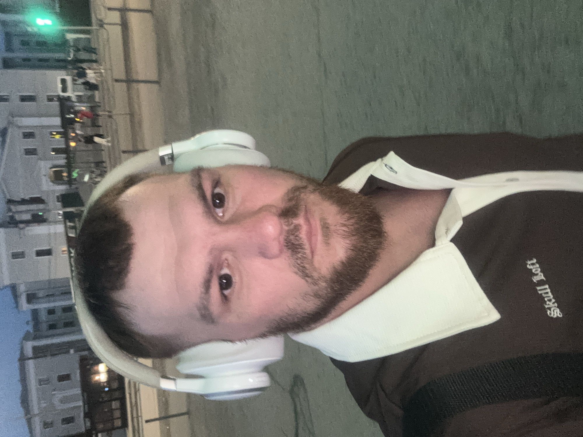 A man is pictured in a selfie outdoors at night, possibly in Perm, Russia, based on the Cyrillic text on his shirt ("Skull Loft"). He is wearing white headphones. He has short, dark hair and a beard. He is wearing a brown shirt with a white collar, with the words "Skull Loft" written in white gothic script on the shirt. In the background, there are buildings and streetlights, suggesting an urban environment.