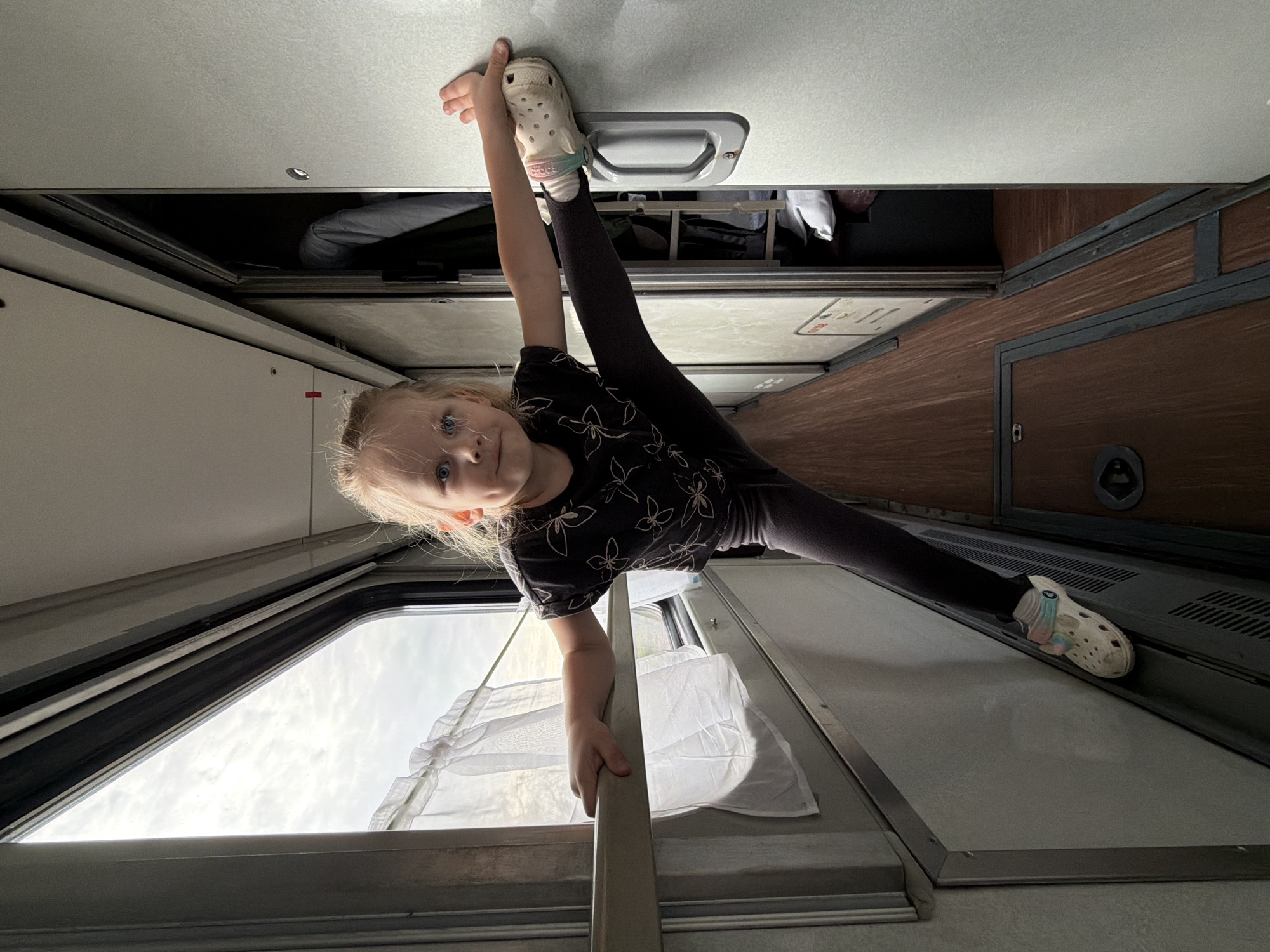 This image captures a young girl performing an acrobatic split within a train compartment in Urusovo, Russia. The photo appears to be taken with the camera rotated, giving the impression that the girl is positioned vertically or upside down.
The main subject is a blonde-haired young girl with blue eyes, looking directly at the camera with a playful and confident expression. She is wearing a dark t-shirt with a light floral pattern and dark leggings. On her feet are light-colored Crocs. She is mid-split, with her left leg extended upwards, her left foot braced against an overhead handle or fixture on the train's ceiling/upper bunk structure. Her right leg extends downwards, and her right hand holds onto a metal railing or support bar for balance.
The setting is the interior of a train carriage, likely a sleeping car or couchette, characterized by functional design and compact spaces. Visible features include light-colored ceiling panels, a window to the left showing an overcast sky, and a white sheer curtain hanging near the window. To the right, there are dark wooden-textured wall panels and what appears to be a closed compartment or door. Above the girl's head, an open overhead storage area reveals some folded fabric or luggage. The lighting suggests it is daytime, with soft, diffused light coming through the window. No discernible text is clearly visible. The overall mood is one of lightheartedness and youthful energy during travel.