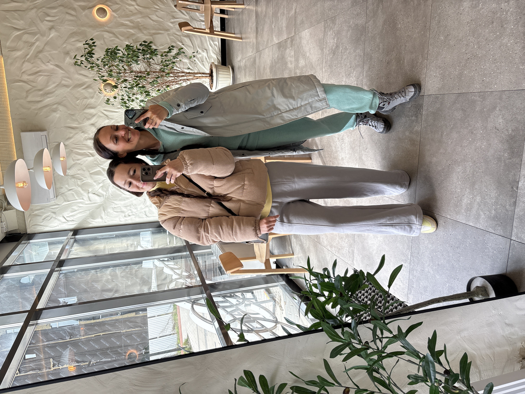 Two women are taking a selfie indoors. The background includes a textured white wall, a faux tree in a white pot, and a wooden airplane toy. One woman wears a beige puffer jacket and gray sweatpants, while the other wears a light gray coat and teal pants. They are in a room with a large window and a view of buildings under construction or renovation. The interior setting appears to be a café or restaurant. While the city is difficult to identify from this photo, based on available information, I am unable to confirm it is in Ulyanovsk, Russia.