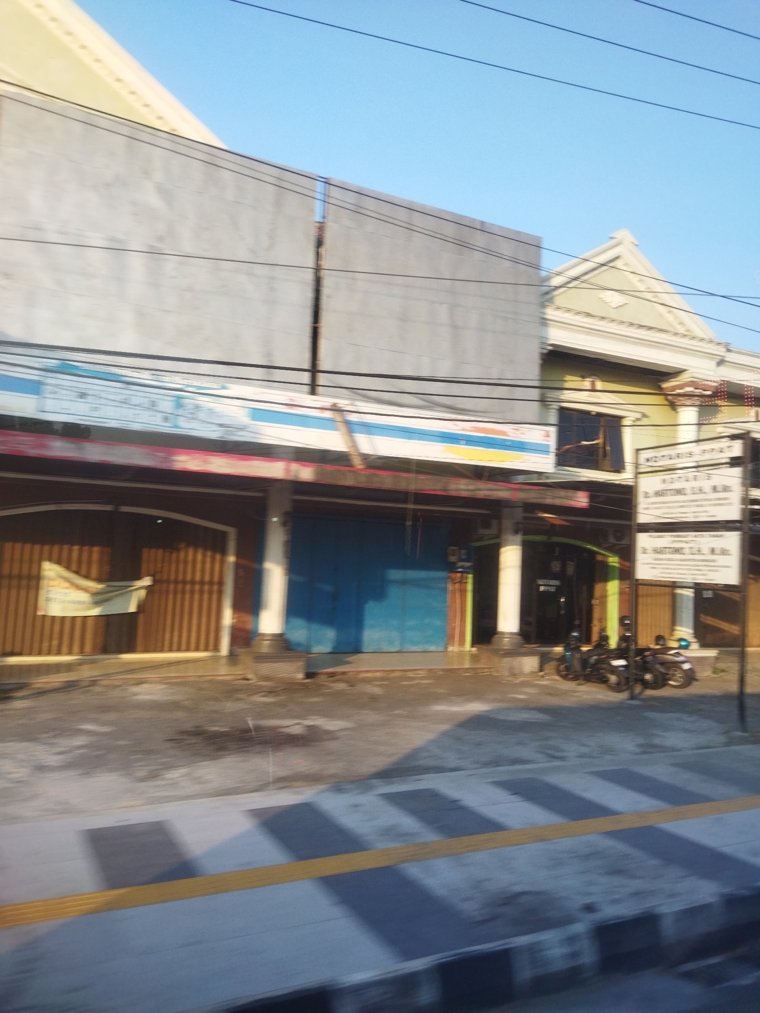 In Wonokarto, Indonesia, a street view captures a commercial building with multiple storefronts under a clear blue sky. The building's facade features a large grey panel on the left, followed by a row of shops with brown wooden roll-up doors and blue metal doors. A prominent blue and white banner stretches across the upper part of the storefronts. To the right, a yellow-green building with an ornate triangular pediment stands adjacent. A black signpost with white text, indicating "NOTARIS & PPAT" and a name, is positioned in front of this building. Parked motorcycles, three in total, are visible on the sidewalk in front of the shops. The sidewalk itself has a textured surface with yellow tactile paving stripes. The overall scene depicts a typical urban commercial area during daylight hours.