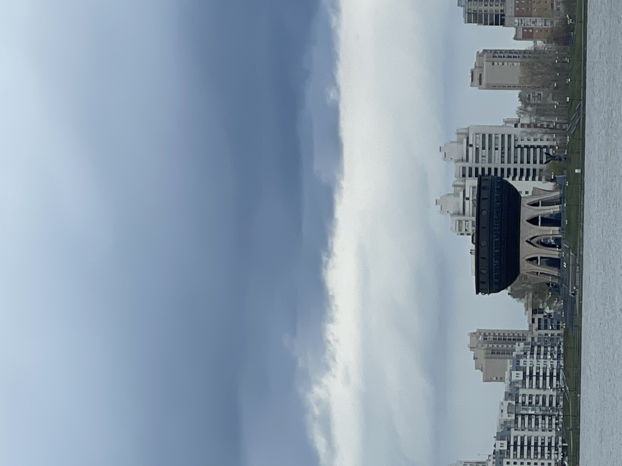 The image, when viewed in its intended landscape orientation, presents a serene urban panorama of Kazan, Russia, along a wide body of water. The upper portion of the frame is dominated by a sky that transitions from a light, hazy blue on the left to a denser expanse of white and grey clouds on the right, indicative of an overcast day.
Along the horizon, a cityscape unfolds, characterized by numerous modern, multi-story buildings, mostly in light tones, featuring repetitive window patterns and balconies. A prominent architectural feature is a large, dark-colored, cylindrical structure with distinct arched openings and an ornate upper rim, situated among the other buildings near the water. This iconic building is the Kazan Family Center (Palace of Marriage). Faint, illegible text, likely Cyrillic script, can be observed along its upper circumference.
A strip of green land, dotted with sparse trees and small streetlights, separates the urban structures from the expansive body of water that occupies the foreground. The water appears calm and reflects the muted light of the cloudy sky. There is no discernible human activity or specific event taking place. The overall impression is one of a quiet, grand urban landscape under a soft, diffused light, consistent with an overcast daytime setting in Kazan.