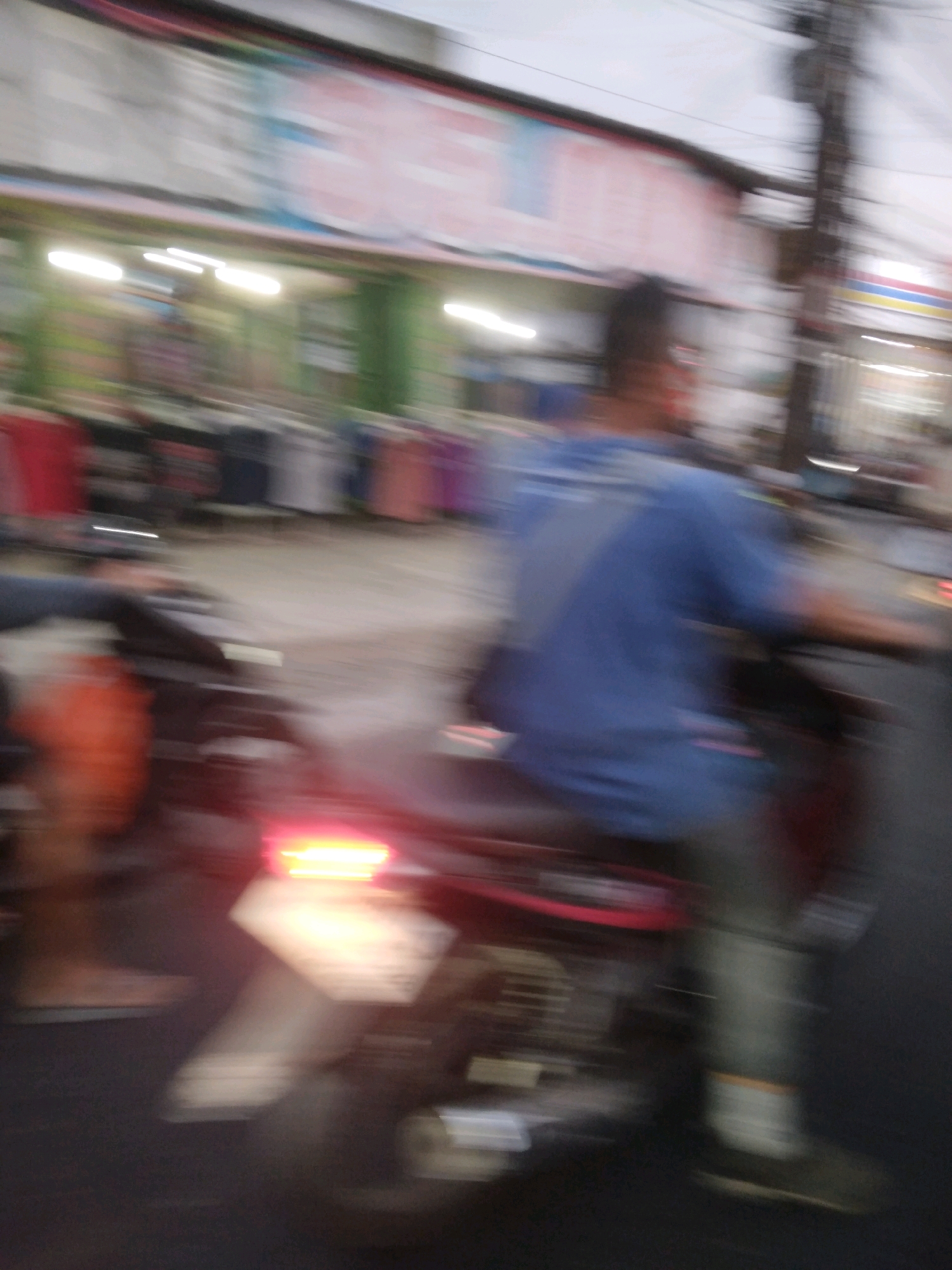The image depicts a blurred motion scene of a person riding a motorcycle in Sokaraja, Indonesia. The rider, wearing a blue shirt and grey pants, is in the foreground, with their body mostly obscured by motion blur. The motorcycle's brake light is illuminated, indicating it is either stopped or decelerating. In the background, a row of shops with their lights on is visible, with clothing racks displaying various garments. The overall impression is that of a busy street scene, possibly during twilight or evening, given the illuminated shop lights and the soft, diffused natural light. The presence of utility poles and overhead wires further suggests an urban environment.
