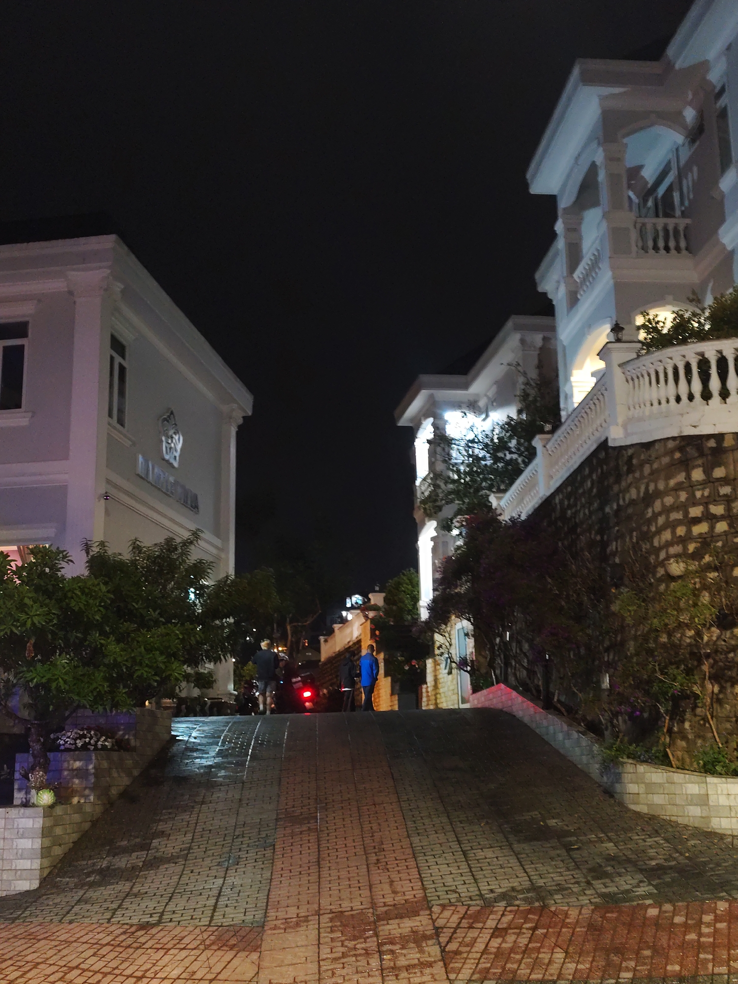 This image shows a view up a sloping street paved with interlocking bricks, bordered by buildings on either side, at night in Da Lat, Vietnam. The building on the left features the sign "DU LICH CENTRO," while the building on the right has a stone wall foundation and a white balcony with balustrades. In the middle distance, there are several people near a vehicle with illuminated red taillights. Street lights are visible in the distance. The sky is dark. Trees and other greenery are planted near the base of both buildings.
