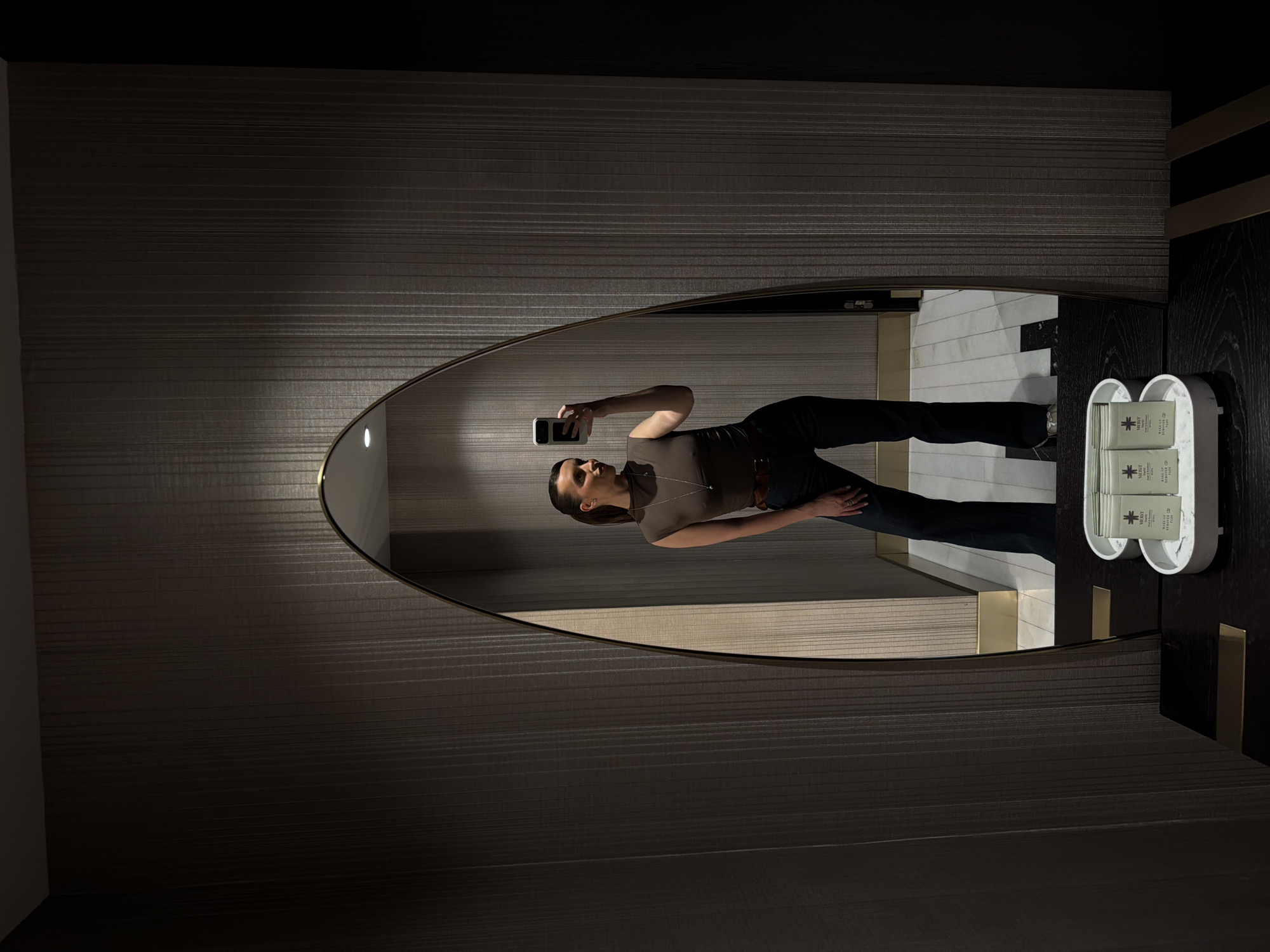 A person is visible within the reflection of an oval mirror. The person is holding a cell phone, seemingly taking a picture. They are wearing a brown top, dark blue jeans and a necklace. The location appears to be a bathroom with vertically striped wallpaper. Next to the mirror is a white container holding small boxes with the word "MERIT" printed on them. The setting could be the Avala Resort & Villas in Budva, Montenegro.