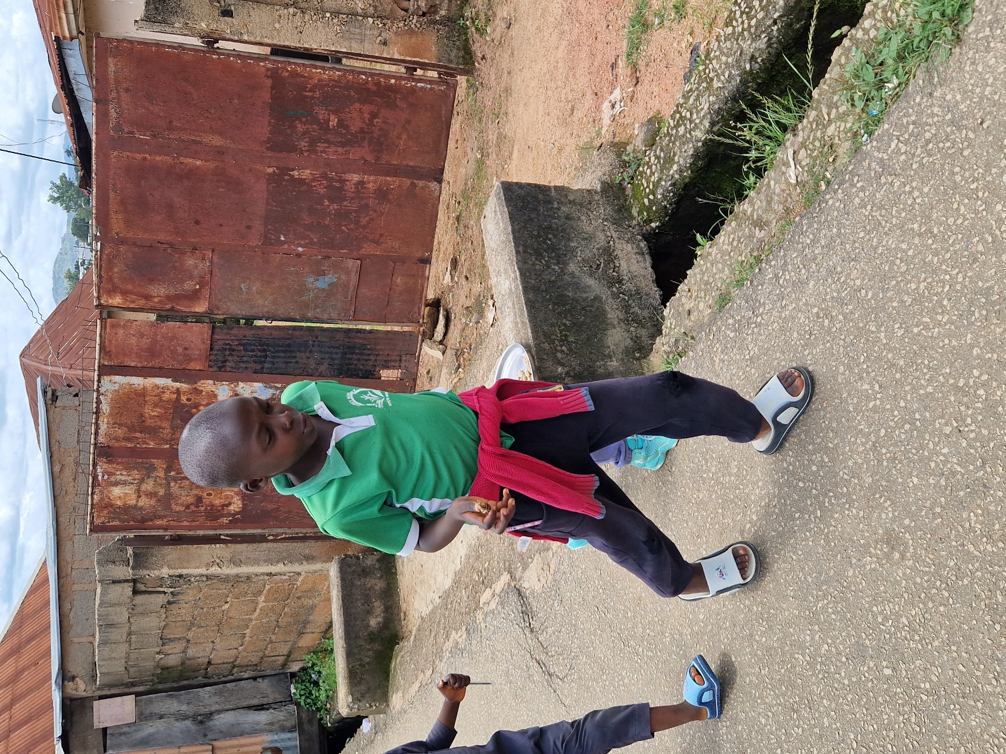 A boy is walking on a paved path in an urban environment. He is wearing a green t-shirt with a white collar and a logo, dark pants, and sandals. A red sweater is tied around his waist. Another child's legs are visible to his left, wearing blue sandals. In the background, there is a rusted metal gate, brick walls, and buildings with corrugated metal roofs. A concrete drain runs along the side of the path. The sky is overcast.