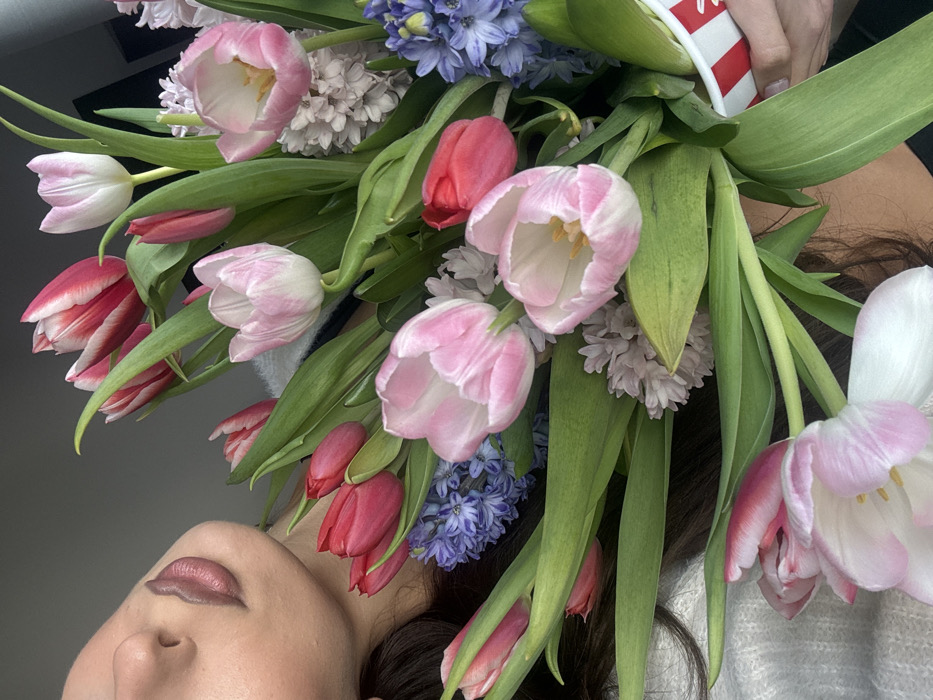 The scene is located in Sofia, Bulgaria, and depicts a close-up, high-angle vertical shot of a large bouquet of fresh flowers held by an individual. The upper left portion of the frame shows the lower half of the individual's face, including the nose and lips, which are coated with a matte nude-toned lipstick. Dark hair cascades around the visible facial features. The individual is wearing a light-colored, possibly white or light grey, textured garment. The central focus is a dense arrangement of spring flowers, primarily tulips and hyacinths. Tulips are present in various colors, including solid pink, pale pink with white streaks, vibrant red, and red with white stripes. Many tulips are partially open, exposing their yellow or white stamens. Clusters of blue and light mauve hyacinths are interspersed throughout the bouquet, adding texture and volume. The flowers are held within a white container adorned with red horizontal stripes, resembling a disposable cup or tub, which is grasped by the individual's right hand visible on the lower right. The background is an out-of-focus, plain grey surface, suggesting an indoor setting.