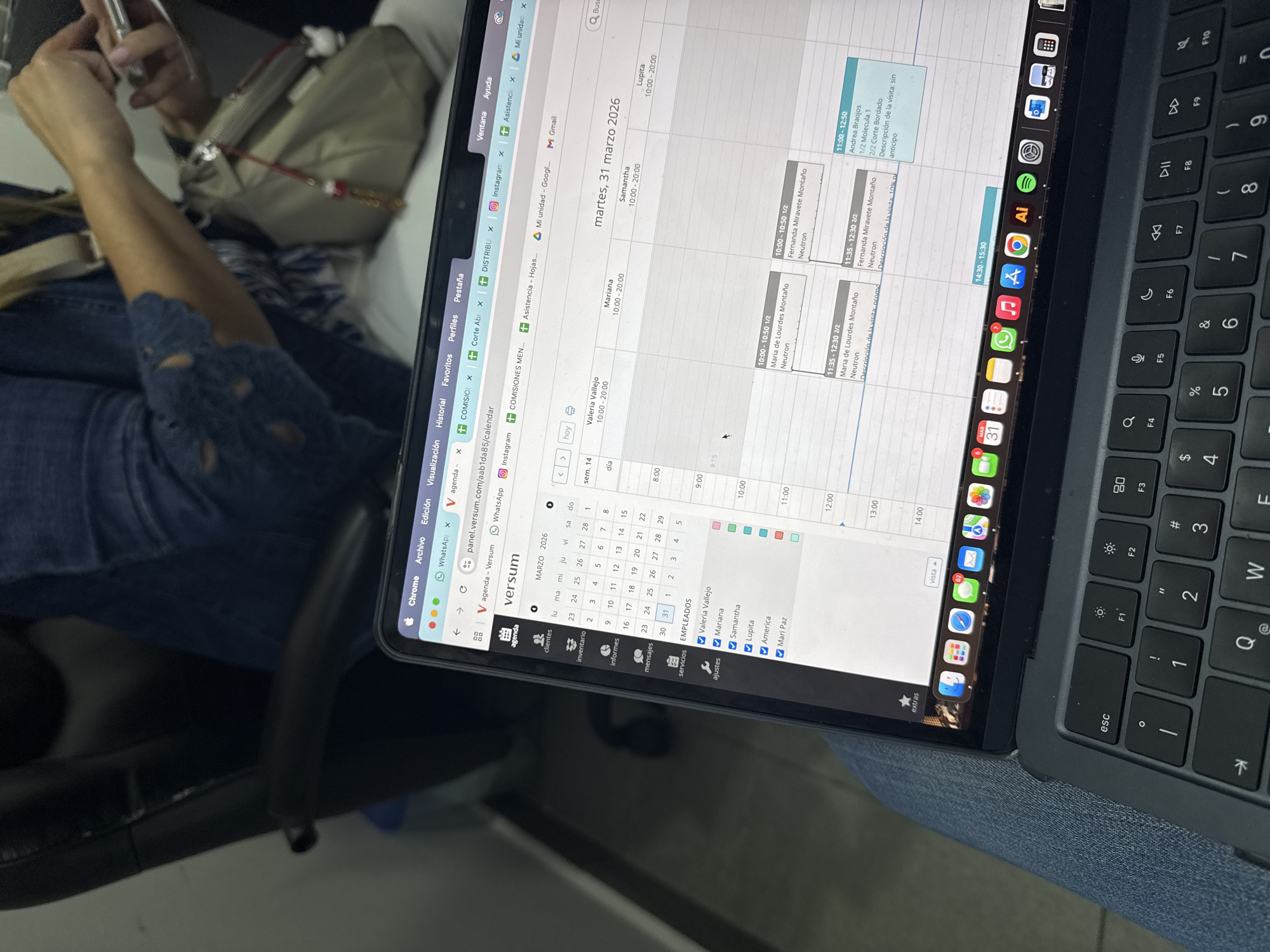 Here is a detailed description of the image:
**Scene Overview:**
The image appears to be taken from a first-person perspective, showing a laptop screen displaying a calendar application. A person is partially visible, sitting in a chair and wearing denim.
**Laptop Screen Details:**
The laptop screen shows a calendar application, likely a business scheduling program. The date displayed is "martes, 31 marzo 2026" (Tuesday, March 31, 2026). The calendar has various entries showing appointments for employees named Valeria Vallejo, Mariana, Samantha, and Lupita, all scheduled from 10:00 to 20:00. Other appointments are visible with specific times and details, such as "Maria de Lourdes Montaño" and "Fernanda Miravete Montaño" with "Neuron" noted alongside them. There is an appointment for "Andrea Braojos" mentioning "Molecula 1" and "Corte Bordado."
**Applications and System Tray:**
The laptop's system tray at the top shows various icons, including Chrome, WhatsApp, Instagram, and other work-related applications. The dock at the bottom includes icons for Finder, Launchpad, App Store, Messages, Mail, Safari, Calendar, WhatsApp, Notes, and Adobe Illustrator. There's a notification badge of 62 on the dock.
**Physical Environment:**
The background shows a glimpse of the person's clothing (denim), and a bag on the side. The environment appears to be indoors, possibly an office or similar setting.
**Location:**
No specific details in the image confirm the location is San Bernardino Tlaxcalancingo, Mexico, or any other location. The location details would require additional information not present in the image.