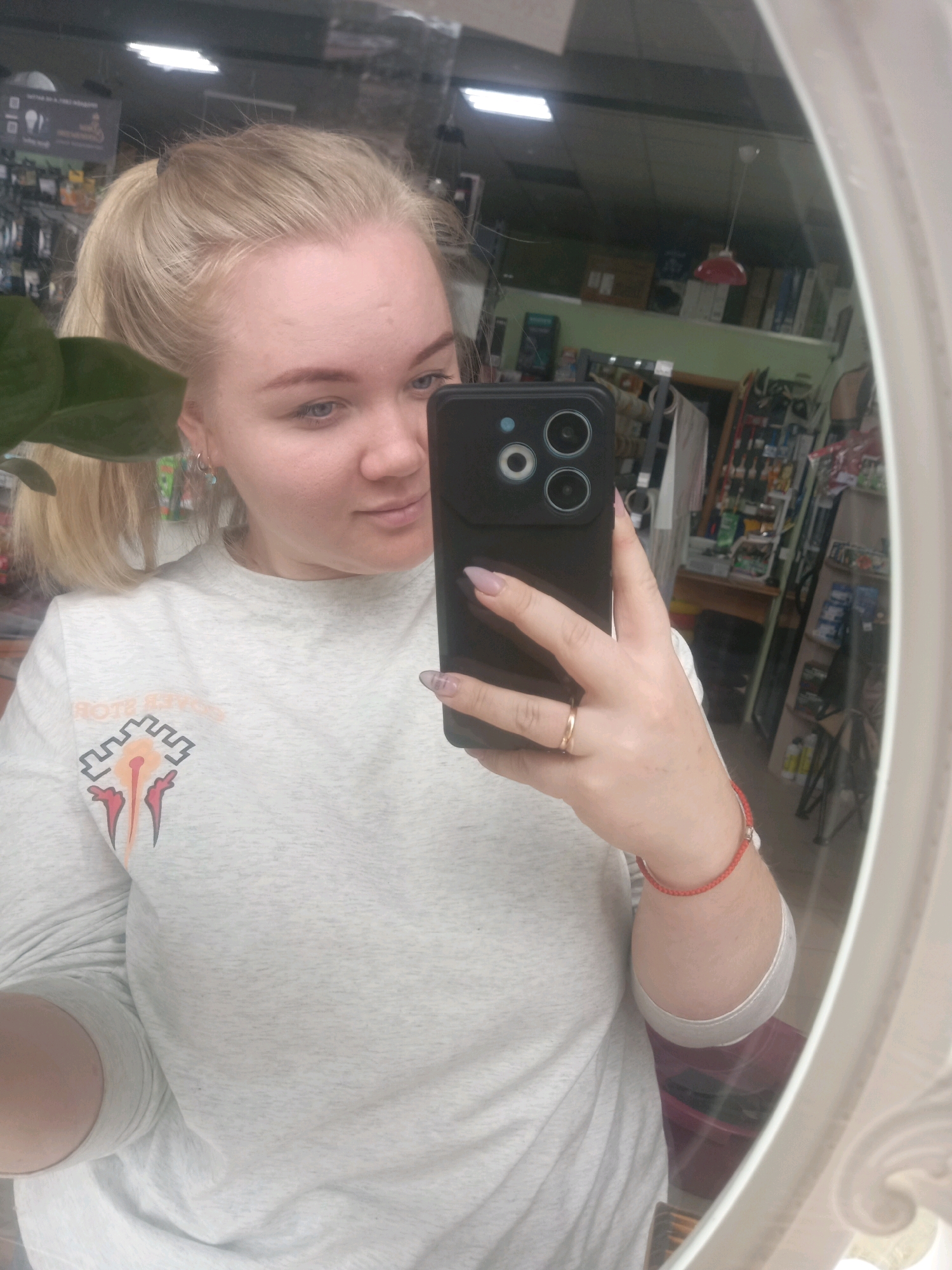 A young woman with blonde hair in a ponytail and blue eyes is taking a mirror selfie in what appears to be a store. She is wearing a light grey long-sleeved shirt with a graphic design on the left chest. She has a gold ring on her right ring finger and a red string bracelet on her right wrist. Her phone is visible, held in her right hand. The background reflects shelves stocked with various items and lighting fixtures, suggesting a retail environment. The overall lighting is adequate for the photograph.