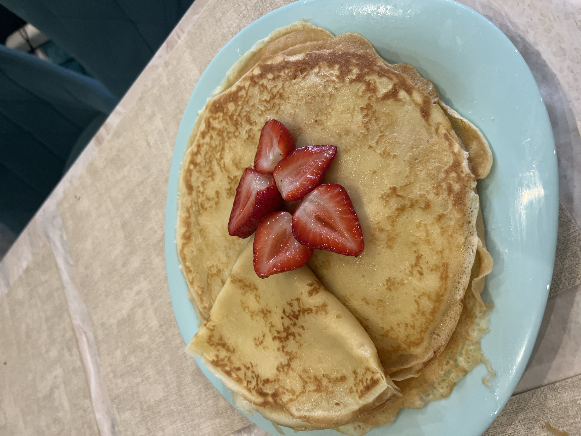 A light blue circular plate is centrally positioned on a beige, textured tabletop surface. The plate holds a stack of thin, golden-brown pancakes or crepes, one of which is folded into a triangular shape and placed atop the stack. Five bright red strawberry slices, appearing fresh and slightly glistening, are arranged across the center of the pancakes. The tabletop surface exhibits a prominent horizontal seam or crease in the upper frame. The blurred background features dark teal upholstered chairs and indistinct lighter objects, indicating an indoor dining environment. The image contains no discernible visual information to identify a specific geographical location within the city of Chaikovsky, Russia, or any other region.
