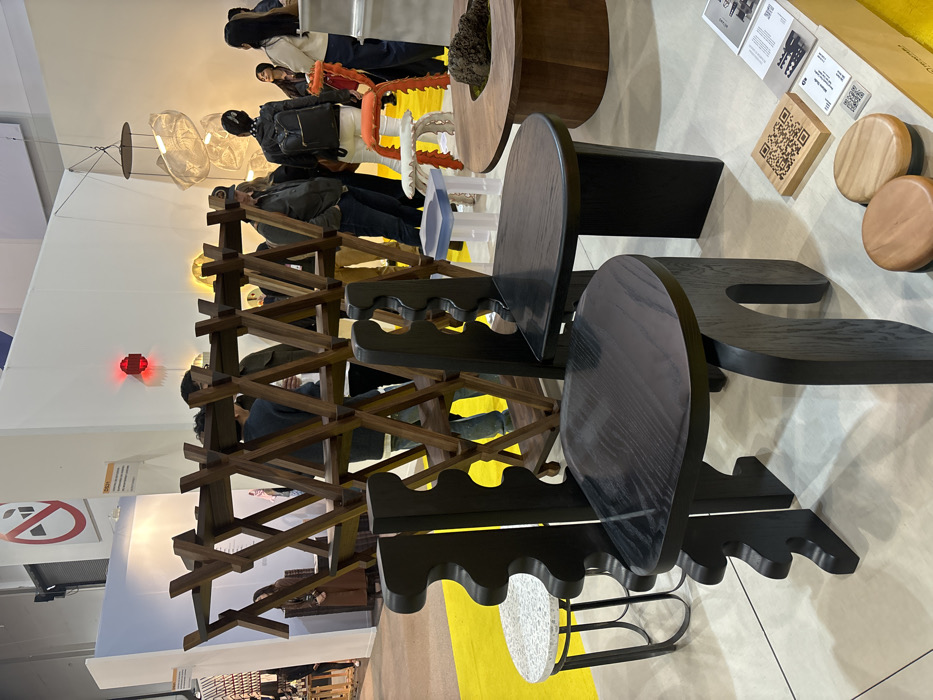 An exhibition space features avant-garde furniture. A slatted wood structure stands tall, while unique black wooden chairs with cutout details are positioned nearby. Round wooden tables and accessories are displayed, alongside signage and promotional materials. People mill about, observing the designs. Overhead, intricate lighting fixtures cast a soft glow. A yellow floor section adds a vibrant touch.