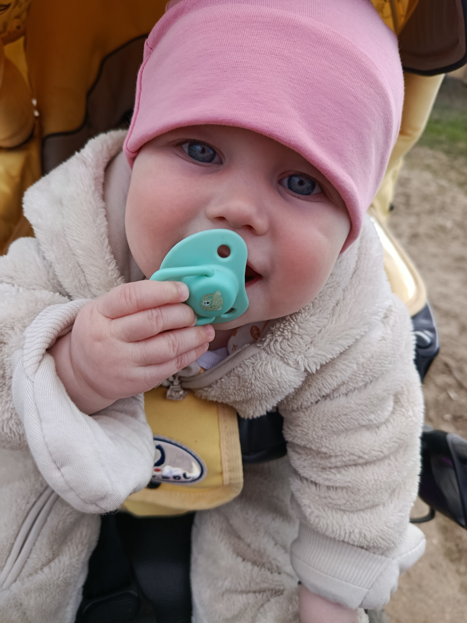 A baby is seated in a yellow stroller, holding a turquoise pacifier with a small green animal design on the front. The baby is wearing a pink hat and a cream-colored, fuzzy hooded jacket. The baby's eyes are blue, and their hand is visible, holding the pacifier. The location is Фоки, Russia.