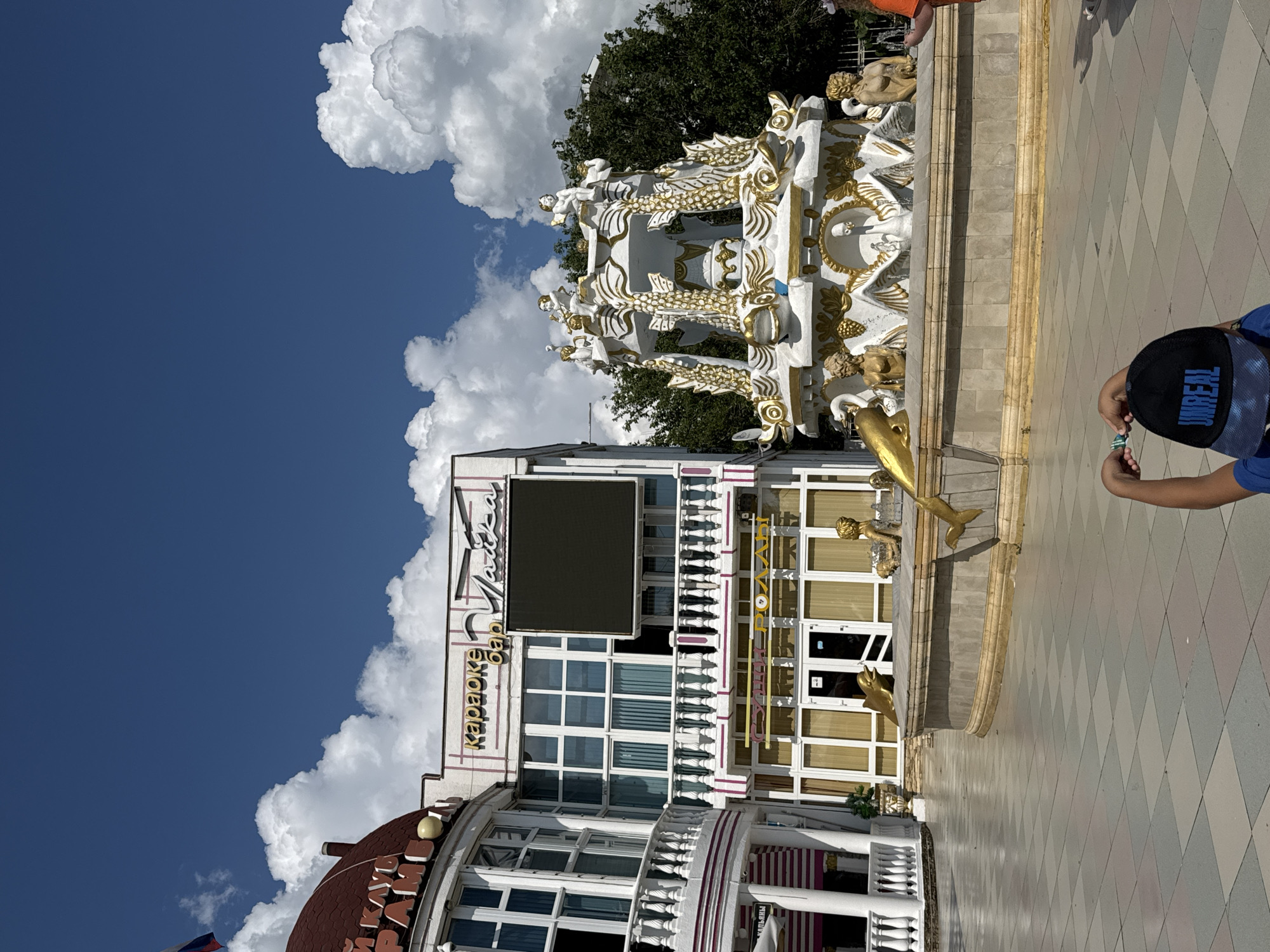 In Myskhako, Russia, on a sunny day with a bright blue sky and scattered white clouds, a vibrant scene unfolds. A grand fountain, adorned with gold accents and featuring sculptures of mermaids, dolphins, and swans, stands prominently in a paved plaza. To the left, a building with a domed roof and a red tiled exterior houses a karaoke bar, with a large black screen mounted on its facade. The building features a white balcony with decorative railings and large windows. Below the karaoke bar, a sign for "Sushi-Rolls" indicates a dining establishment. In the foreground, a child wearing a blue shirt and a black baseball cap with the word "UNREAL" on it is partially visible, holding a small green object. The plaza is laid with a checkered pattern of light and dark grey tiles.