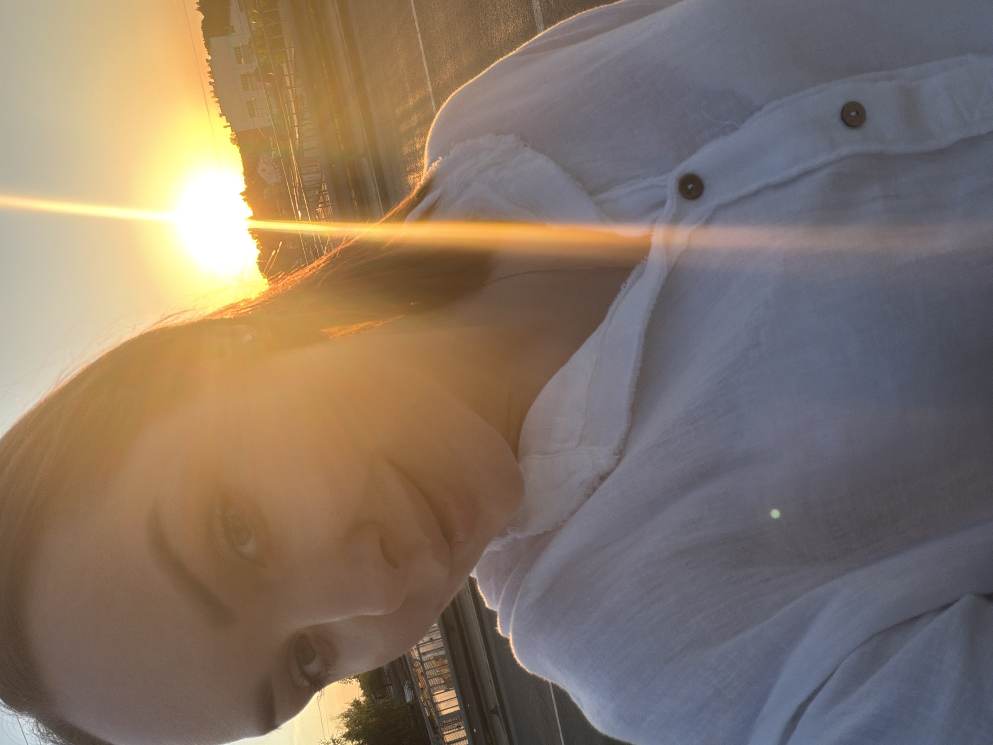 The image is a close-up selfie of a person wearing a white, button-down shirt. The person's face is partially illuminated by a bright light, likely the sun, which is visible in the background. The setting appears to be outdoors, possibly a bridge or elevated structure, with glimpses of buildings and trees silhouetted against the bright sky. The location is plausibly in the city of Chaikovsky, Russia. The primary subjects are the person and the sun, with the action being the taking of a selfie.