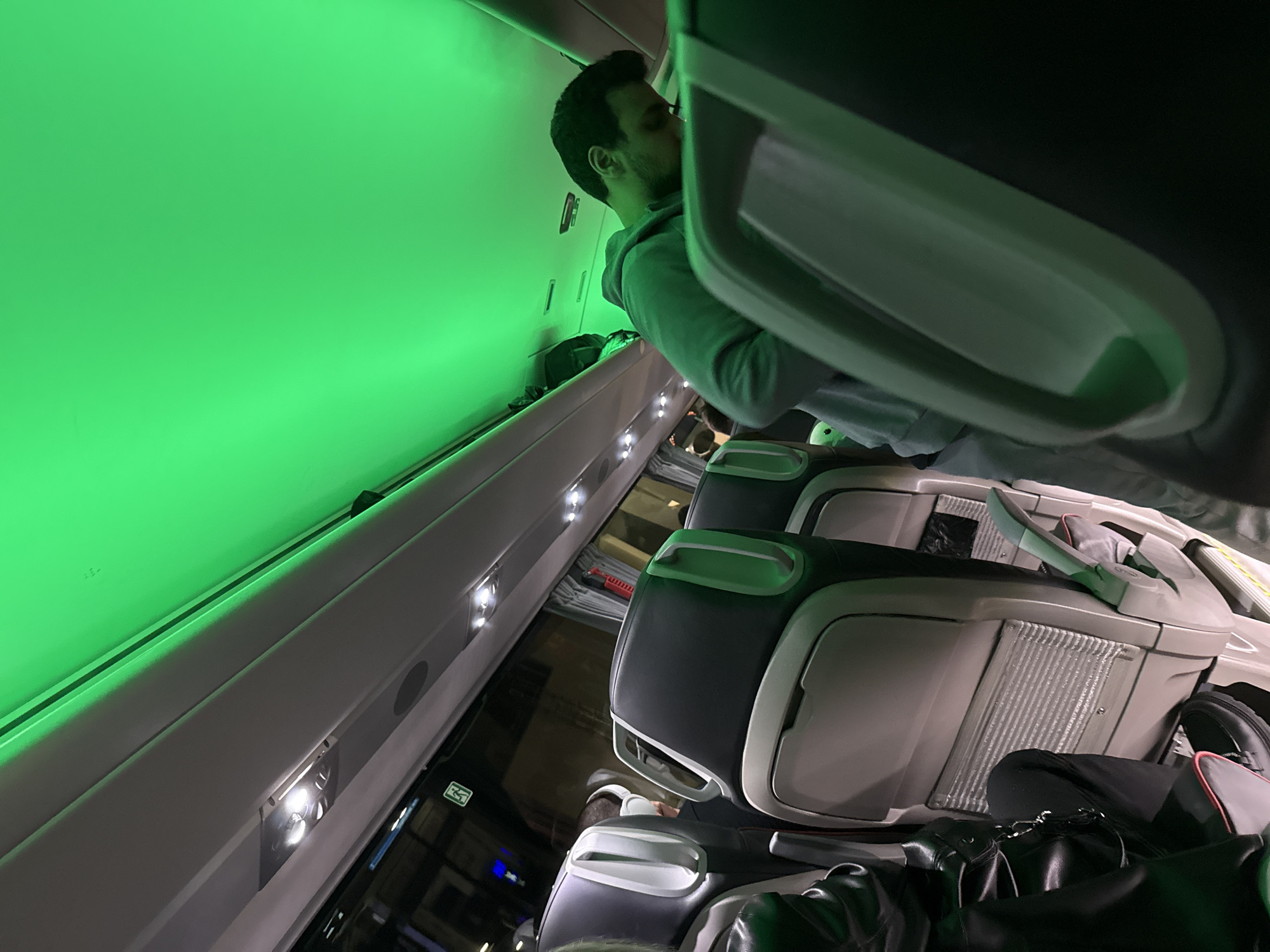 Aboard an airplane, a man sits in his seat, illuminated by green ambient lighting. The interior cabin features overhead lights and signs. Passengers' personal belongings are stowed in the overhead compartment and under the seats. The airplane may be in Varna, Bulgaria.
