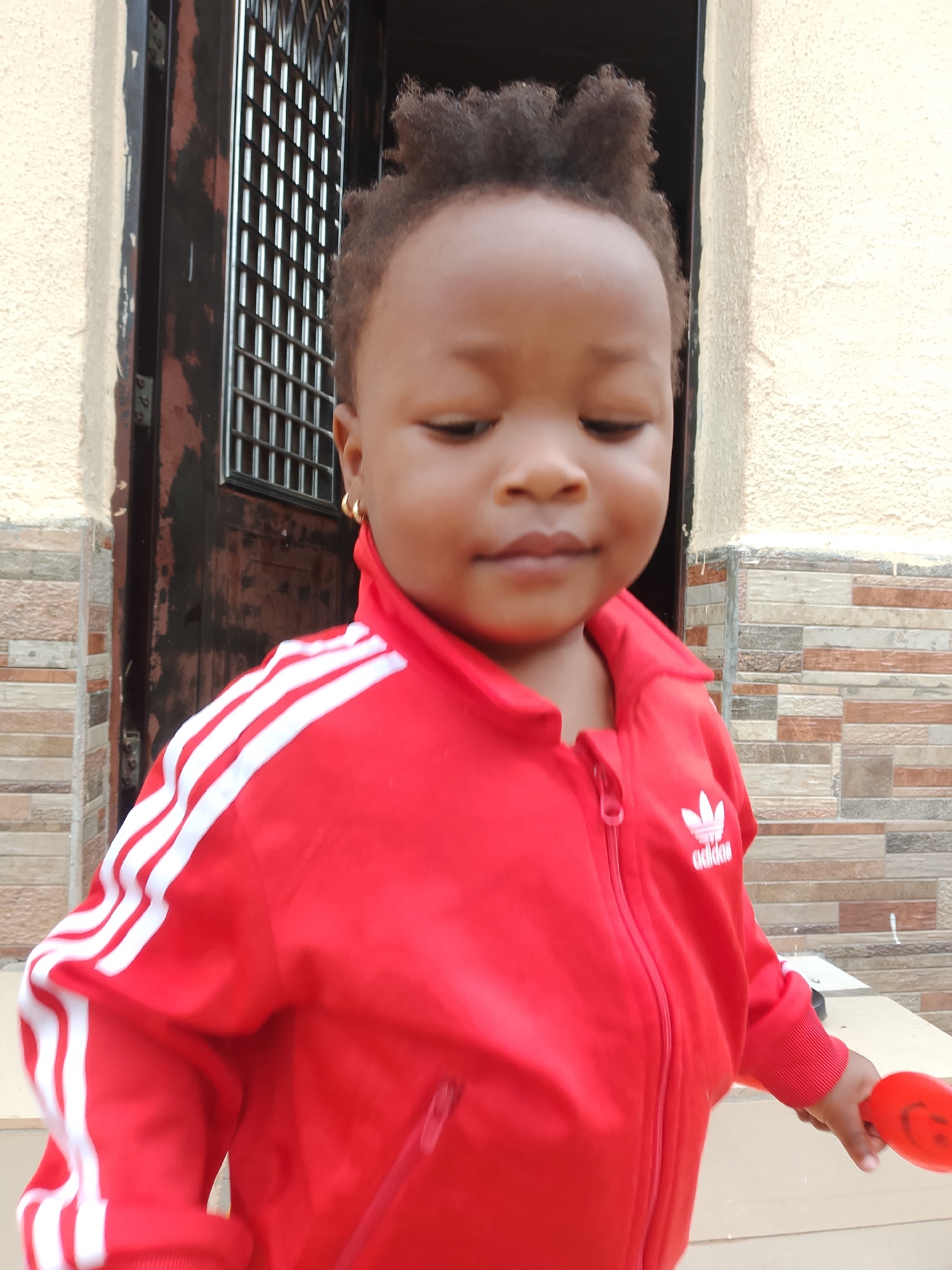 An outdoor, medium-shot, eye-level, slightly low-angle photograph shows a toddler with dark curly hair, wearing a red Adidas tracksuit with white stripes on the sleeves, and a gold hoop earring in her left ear. The toddler is looking down with her eyes closed and a slight smile on her face. She is standing in front of a building with light-colored stucco walls and a brick accent wall. To the left of the toddler is a dark brown door with a metal grate. The toddler is holding a red toy in her right hand. The lighting is natural and bright, suggesting it is daytime. The background is slightly out of focus, with the toddler being the primary subject. The overall scene appears to be residential.