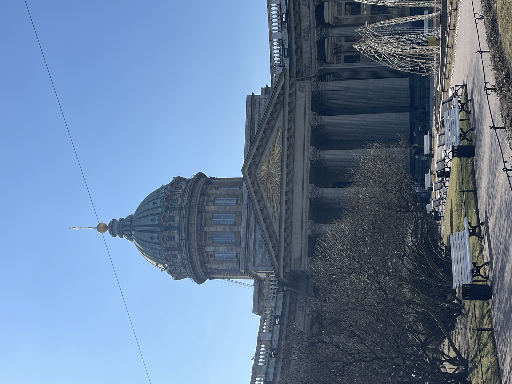 The image shows the Kazan Cathedral in Saint Petersburg, Russia, viewed from a ground-level perspective. The cathedral's dome, adorned with a golden spire, dominates the left side of the image against a clear, blue sky. The architectural details of the cathedral's facade, including columns and ornate carvings, are visible on the right. A tree with bare branches is present in the foreground. A pathway with benches alongside the cathedral is also visible on the right side.