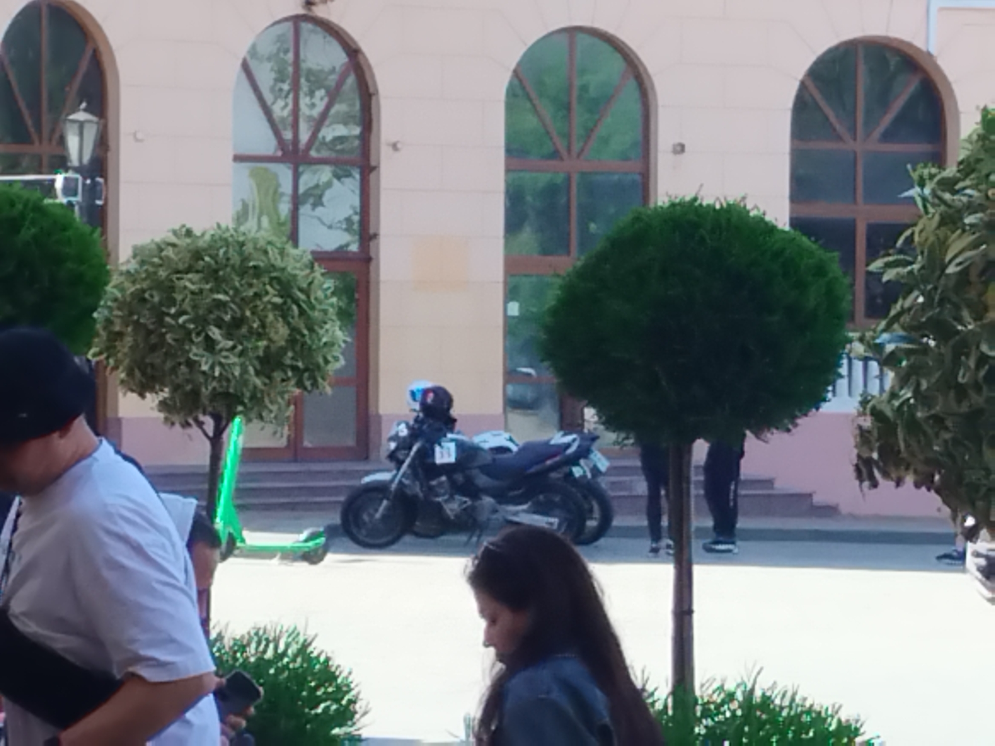 The image depicts an outdoor scene likely in a city setting, possibly Odesa, Ukraine given the architectural style. The background consists of a light-colored building with arched windows and a few decorative elements. A motorcycle is parked on a pedestrian area in front of the building, with a helmet resting on the seat. A green electric scooter is visible near the motorcycle. Several people are present, including a person wearing a light-colored t-shirt and a black cap who is partially visible on the left. A girl with long dark hair is walking in the foreground. Two people are standing by the stairs near the motorcycle. The scene includes several shaped topiary trees. The environment suggests a public area, possibly a plaza or a street with pedestrian access.