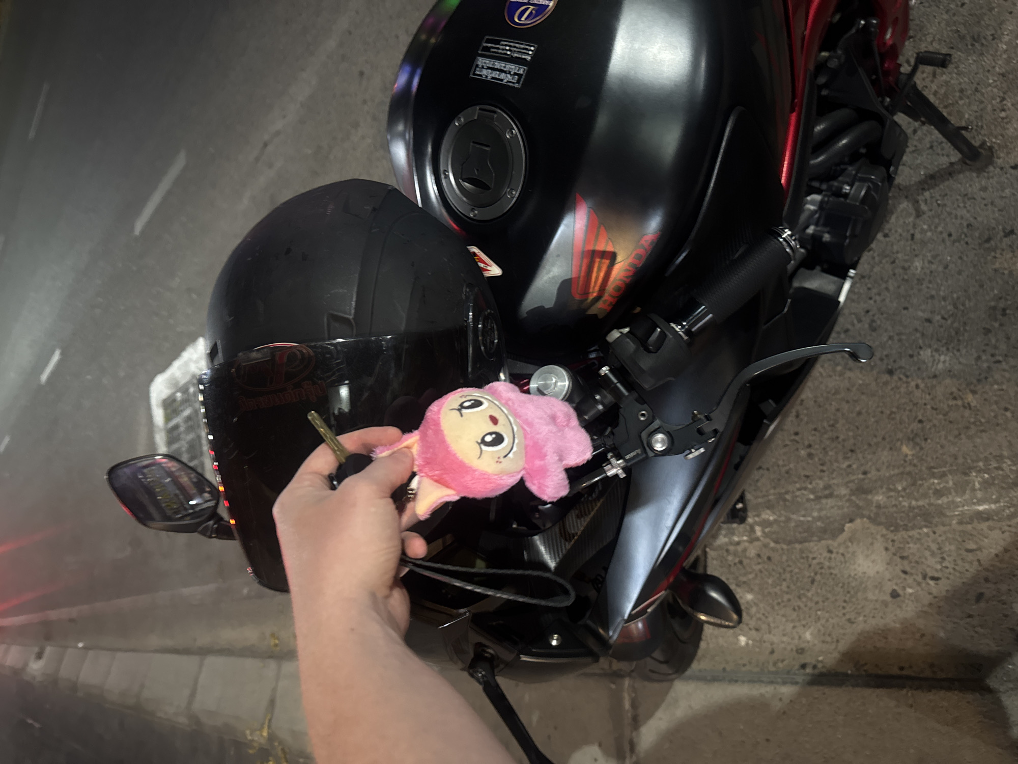 A person's hand holds motorcycle keys and a pink, plush doll keychain near the handlebars of a black and red sport motorcycle at night. A black full-face helmet rests on the motorcycle's matte black fuel tank. The motorcycle is parked outdoors on a paved surface, likely a street or parking lot, with a concrete curb visible on the left side.
The motorcycle features a matte black fuel tank with red "HONDA" lettering and accents on its side. A circular fuel cap is centrally located on the tank. Above the fuel cap, a blue emblem sticker is visible, along with multiple lines of smaller white text below it, possibly warning labels or specifications. Engine components and a kickstand are visible on the right side of the motorcycle. The left rearview mirror reflects the dark surroundings.
The keys, held by a light-skinned hand, have a brass-colored blade. Attached to the keys is a fluffy pink keychain resembling a doll with a round head and large, dark eyes. The scene occurs at nighttime, indicated by the overall darkness and reflections of artificial lights. The ground appears somewhat damp. This urban setting is consistent with a location such as Pattaya City, Thailand.