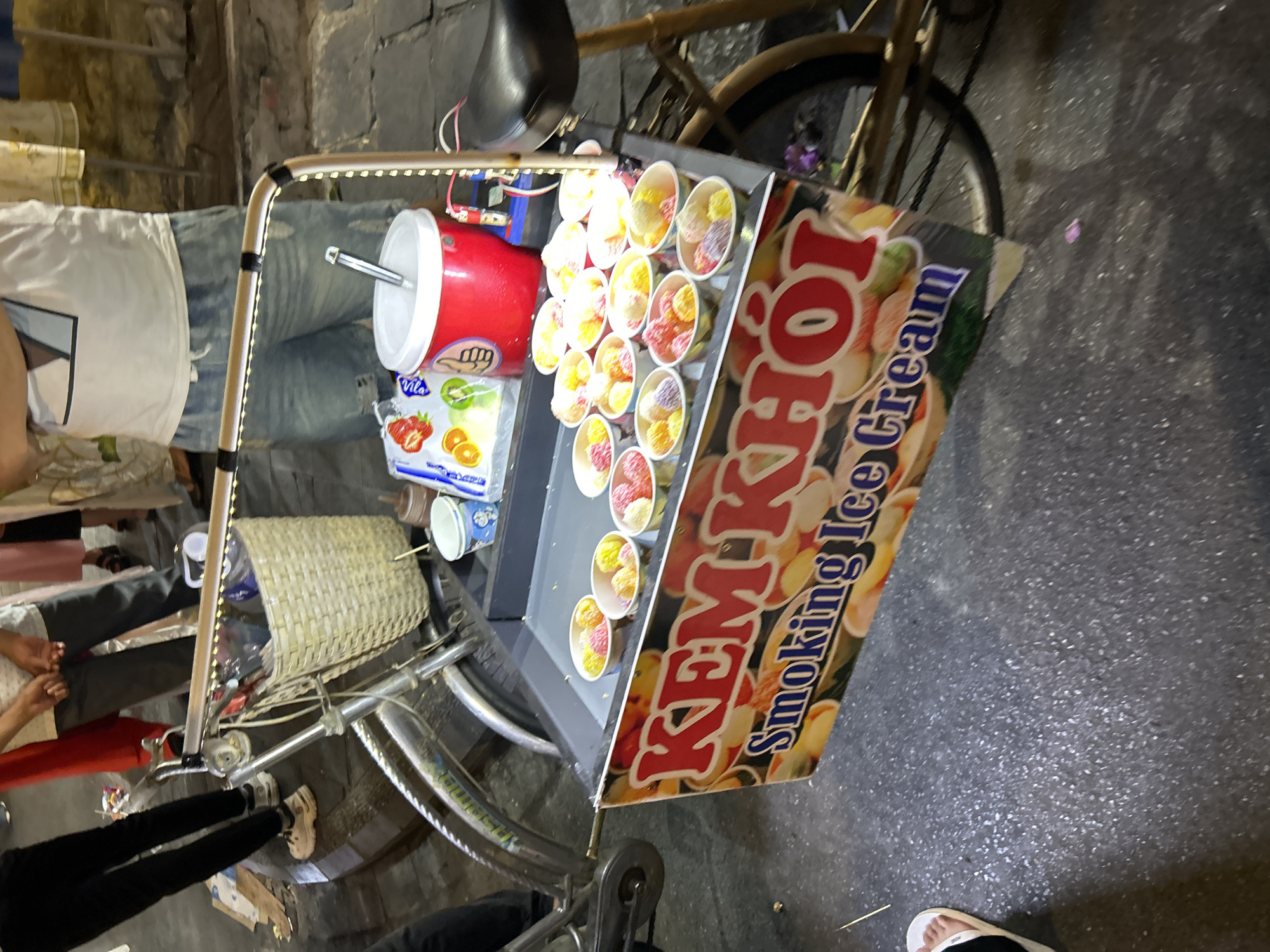 The image depicts a street vendor selling ice cream from a bicycle cart in Hội An Ward, Vietnam. The cart is adorned with a colorful banner displaying the words "KEM KHOI Smoking Ice Cream". A variety of ice cream cups, filled with colorful scoops, are arranged on the cart. Behind the cart, a person in a white t-shirt and denim shorts stands near a large red container. Other individuals, partially visible, are seen in the background, suggesting a bustling street environment. The ground is paved with asphalt.