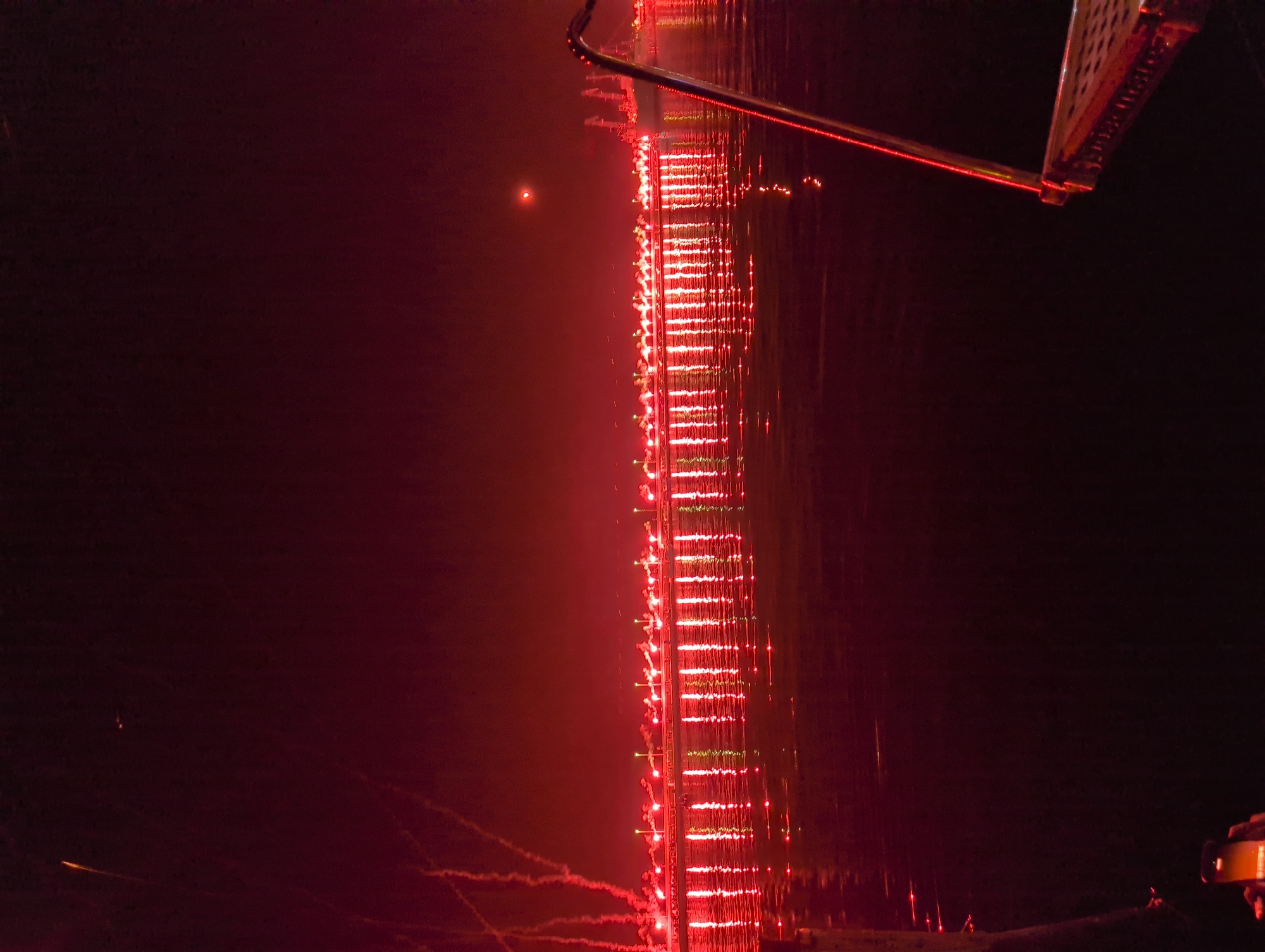 The image captures a waterfront scene at night, likely in the city of Rijeka, Croatia, given the presence of maritime infrastructure. The dominant color is a deep red, cast by a display of fireworks or light show, which illuminates a long line of what appear to be ships or boats docked along a harbor or waterfront. These vessels are adorned with numerous bright red lights, creating a continuous illuminated band across the scene.
The water in the foreground reflects these red lights, creating shimmering patterns on the dark surface. In the upper portion of the image, faint trails of red smoke or light from fireworks are visible against the dark sky. The sky itself is a deep, dark red, with a small, indistinct light source, possibly the moon or a distant light, visible near the horizon.
On the right side of the frame, a portion of a metal railing or structure, possibly from a boat or pier, is visible, also reflecting the red light. In the lower left corner, a hand is holding a smartphone, likely capturing the event, with the phone's screen illuminated. The overall atmosphere is one of a festive nighttime event taking place at a coastal location, with the city's port facilities serving as a backdrop.