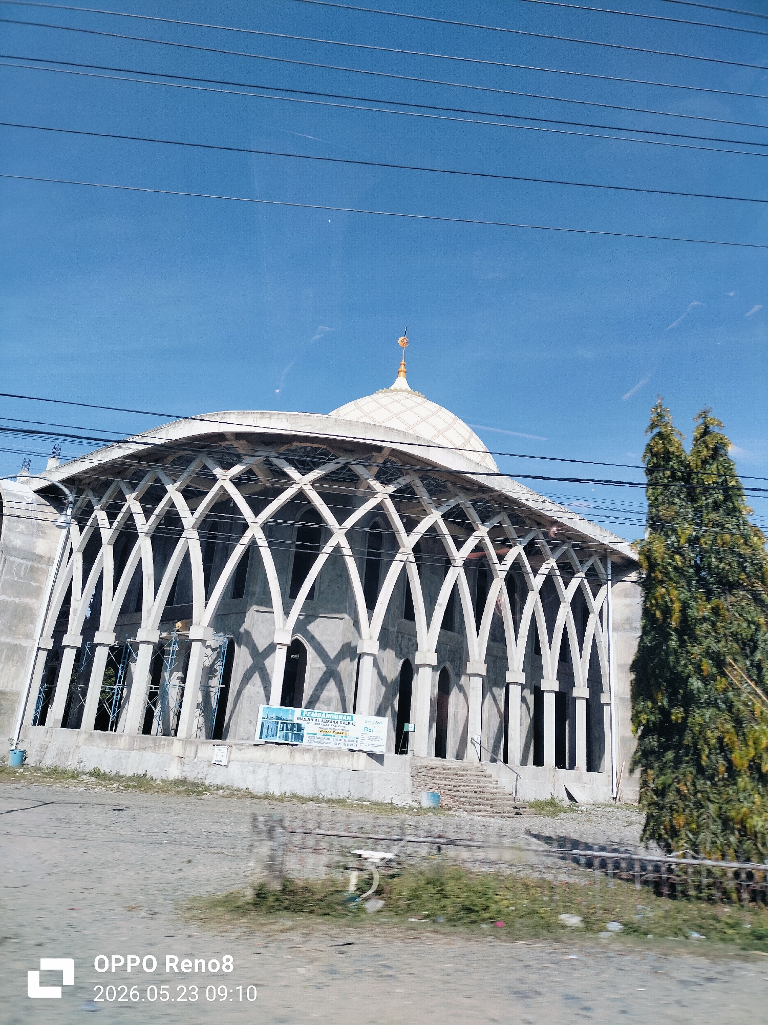 An eye-level shot captures a mosque under construction in Teungoh Baro, Indonesia. The structure features a white dome with a spire, supported by a series of arched white concrete frames. A tree stands to the right of the mosque. The building is surrounded by what appears to be a construction zone. Overhead power lines cross the sky. The sky is blue with some light clouds. A sign is posted at the base of the building. The image was taken on May 23, 2026, at 09:10 AM.