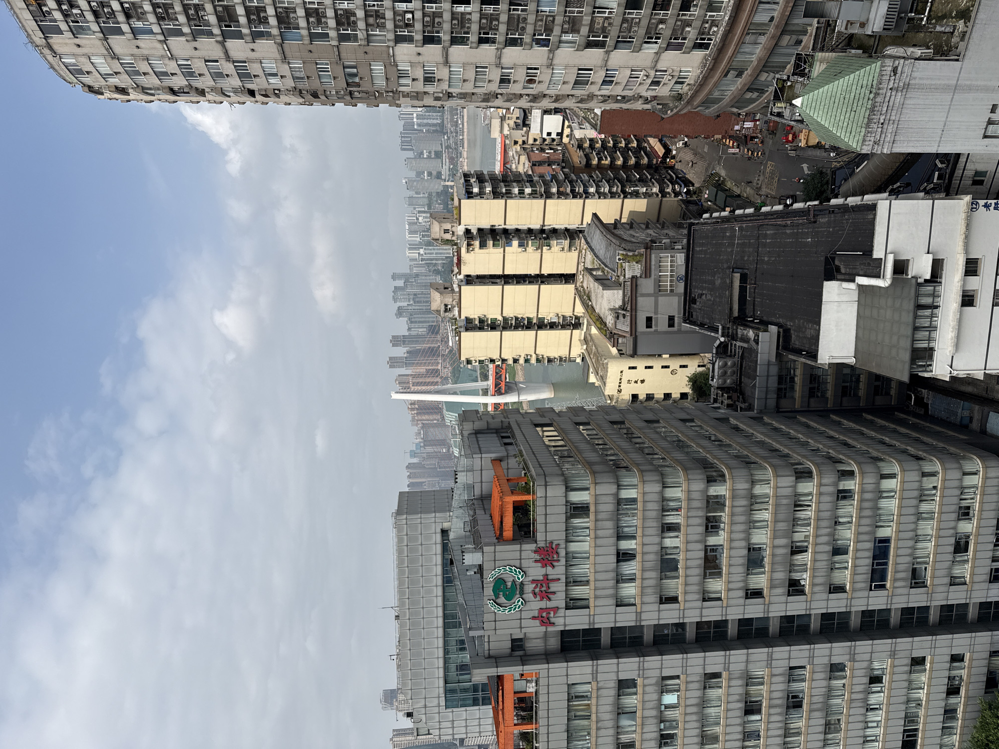 An elevated view in Yuzhong District, Chongqing, China. Tall buildings of concrete and glass frame the view. Other buildings appear to be residential. The sky is partly cloudy. A bridge can be seen in the distance.