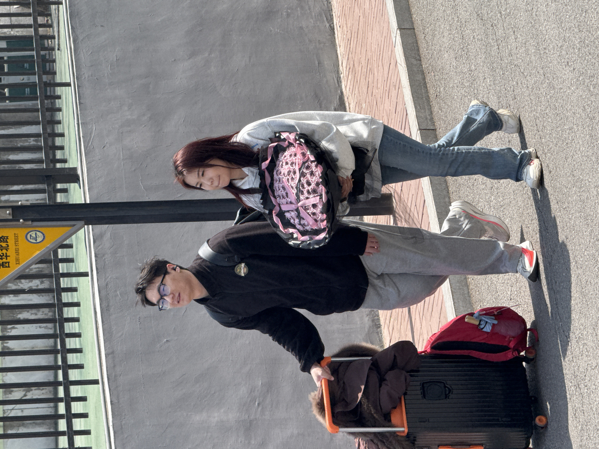 A woman and a man are outdoors next to a street sign for Xihuaibei Road, in what is possibly Zhongtun, China. The man is wearing glasses, a black top, and gray pants; he is also pulling a black suitcase with an orange handle, on which a brown garment and a red backpack sit. The woman, who has long, red-brown hair, is wearing a gray hoodie and blue jeans and carrying a large bouquet of pink roses wrapped in black fabric. They are standing on a gray sidewalk next to a gray wall and a railing.