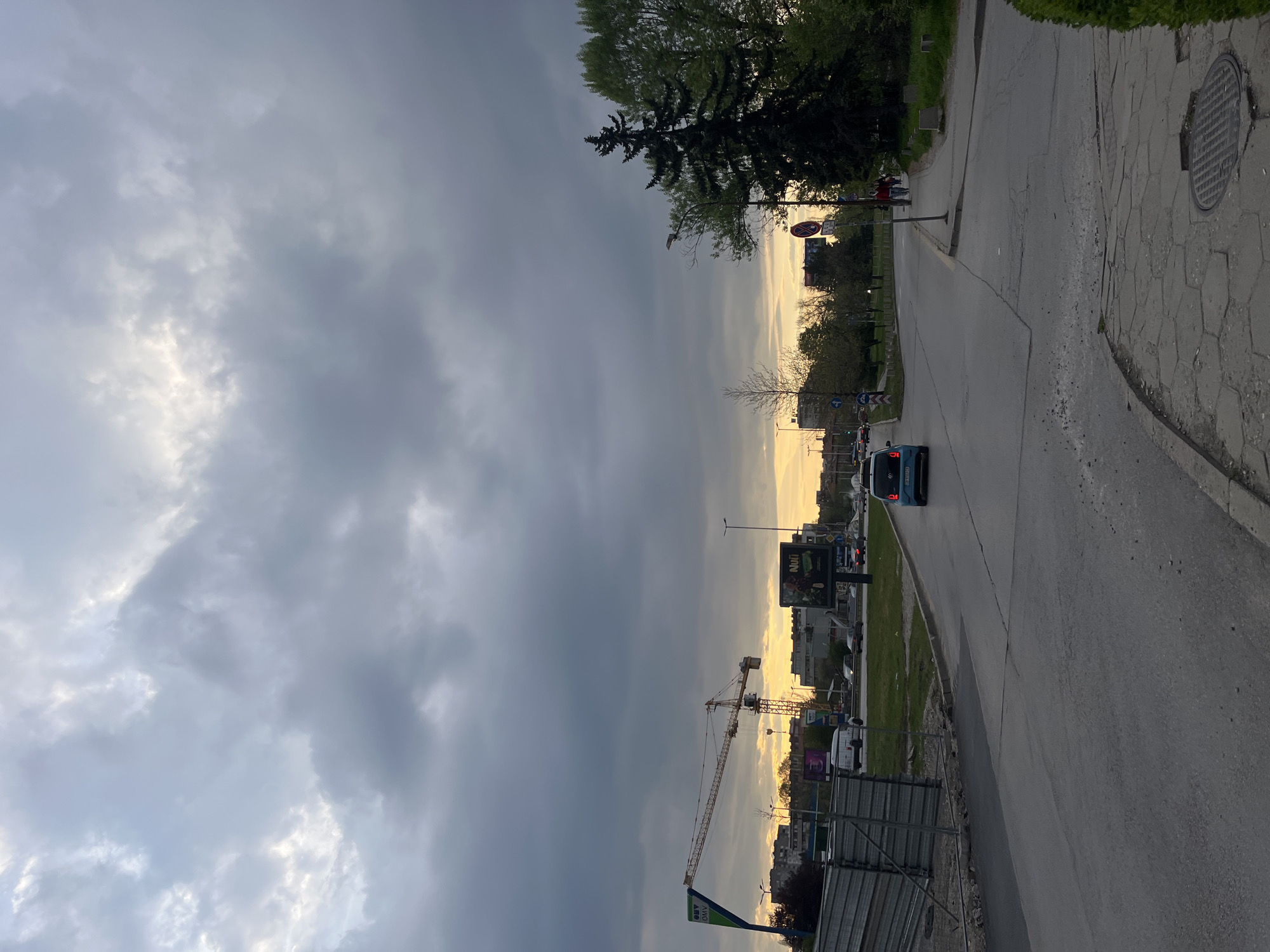 This image presents an urban street scene under a dramatic, cloudy sky, likely captured during late afternoon or early evening. The upper and left portions of the sky are dominated by heavy, dark grey clouds, while a distinct band of golden, yellowish light illuminates the horizon on the right, suggesting a setting sun or a clear break in the clouds.
A multi-lane asphalt road extends from the foreground into the distance, with a few vehicles, including a blue car and a larger blue and white bus or van, indicating light traffic. To the right of the road, a cracked concrete sidewalk features a prominent manhole cover. Lush green grass and various trees, including large, dark conifers and smaller, deciduous trees, line both sides of the road and separate it from the surrounding urban environment.
In the mid-ground, a tall yellow construction crane stands out against the brighter horizon, pointing to ongoing development. Several urban buildings are visible in the background. Along the roadside, multiple billboards are present, one clearly displaying "NIVI" and another partially showing "OMV." Various street signs are also visible, including a red circular prohibition sign and a blue directional sign. A person wearing a red top can be seen walking on the distant sidewalk to the right.
The scene captures a typical city environment in Sofia, Bulgaria, characterized by a mix of infrastructure, natural elements, and signs of construction, all under a striking and atmospheric sky.