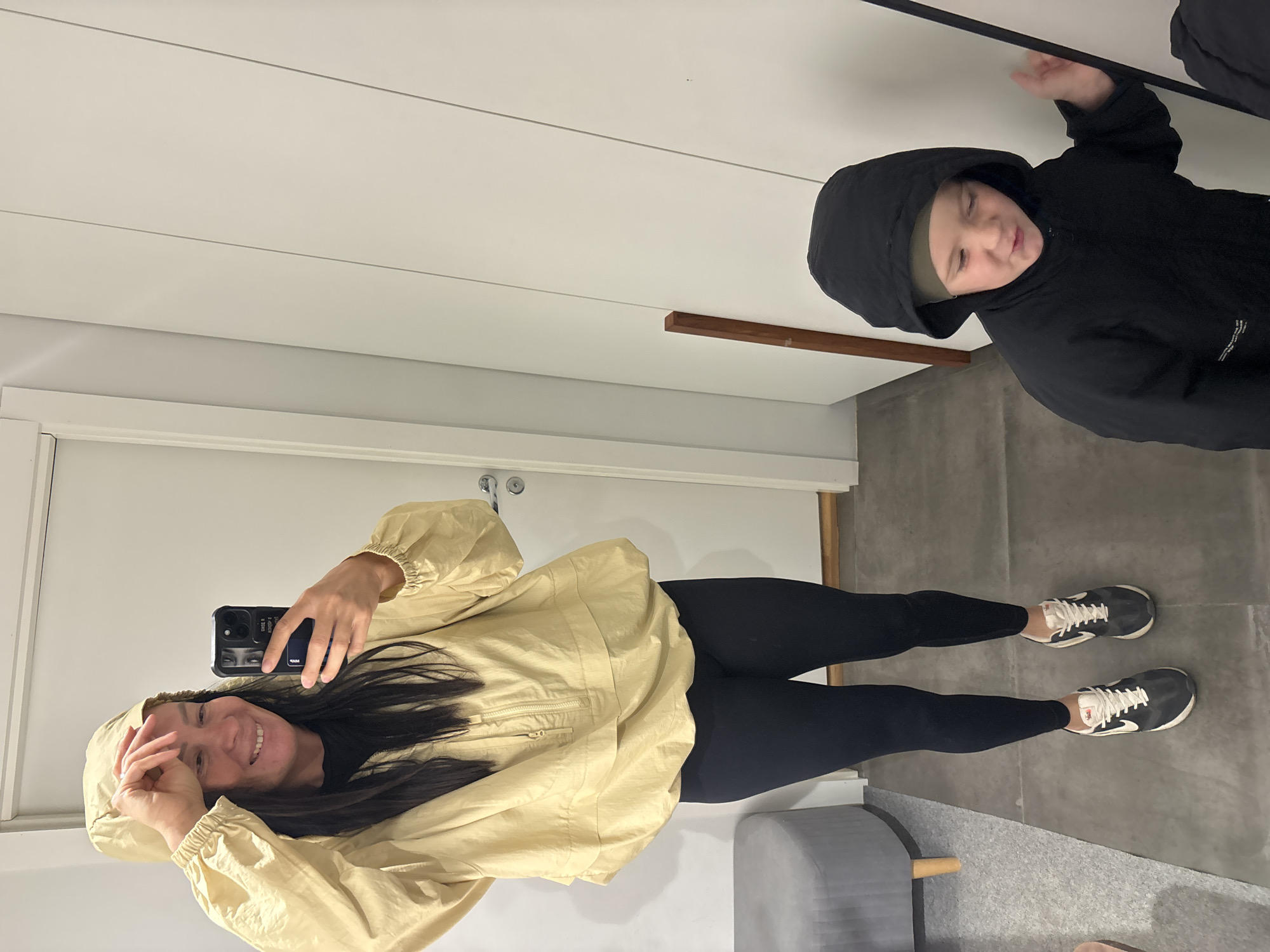 A woman is taking a selfie with a child in a hallway. The woman is wearing a beige hooded jacket and black leggings, with sneakers. The child is wearing a black hooded jacket. The location appears to be an interior space with a white door and gray flooring. The location is likely in Chaikovsky, Russia.