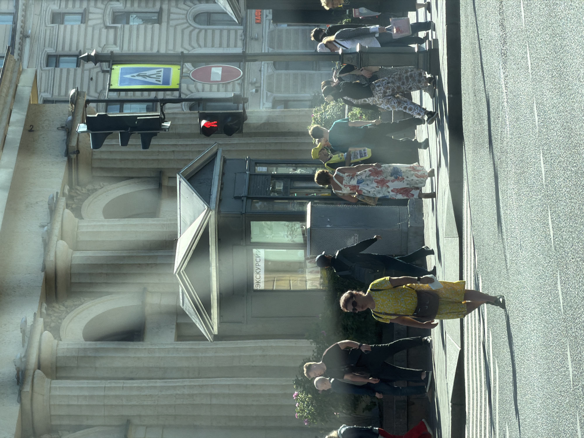 The image captures a street scene in Saint Petersburg, Russia, on a sunny day, likely in the historic center given the architectural style. In the foreground, a pedestrian crossing is visible with a woman in a yellow floral dress walking across it from left to right. She wears dark shoes and carries a brown messenger bag over her right shoulder. The street is paved with asphalt, and sunlight casts long shadows.
Behind the crossing, several people are walking on the sidewalk. To the left, a man in a dark t-shirt and black pants walks away from the camera, carrying a black bag. To his right, another man in dark clothing walks towards the camera. Further back, a kiosk with the word "ЭКСКУРСИИ" (Excursions) written on it is situated.
A traffic light with a red pedestrian signal (a standing figure) is prominently displayed. Adjacent to the traffic light is a pedestrian crossing sign depicting a white figure on a blue background within a yellow border. A red circular sign with a white horizontal bar is also visible below the pedestrian crossing sign.
In the background, a grand building with large columns and arched windows stands. Another building, appearing to be a multi-story structure with visible windows, is to the right. The overall atmosphere is one of a bustling urban environment with people going about their day.