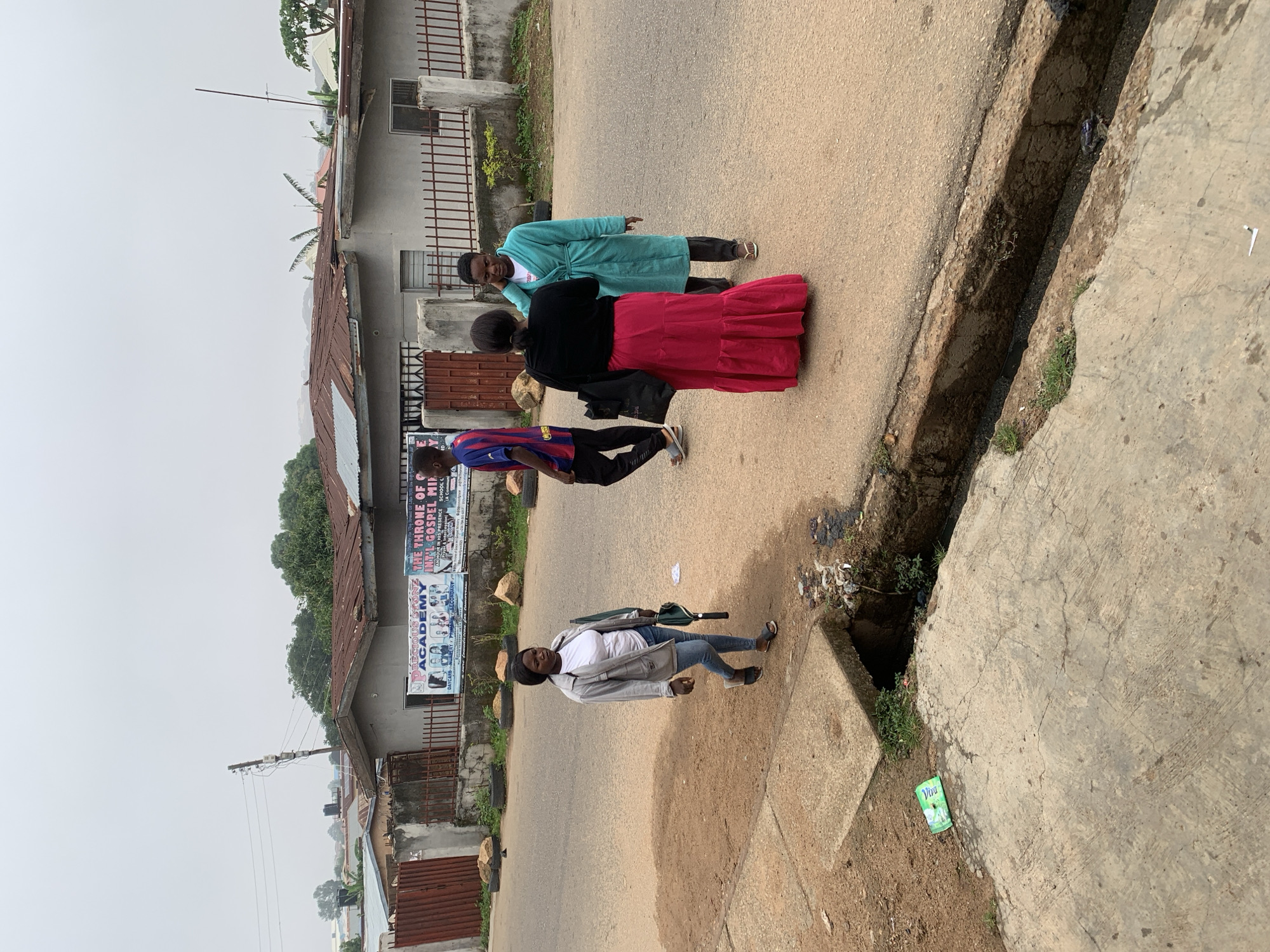 In Jos, Nigeria, on a cloudy day, four individuals are visible on a street. To the left, a woman walks on a concrete path beside a dusty road, carrying a green umbrella and a handbag. She is wearing a white t-shirt and a grey jacket with blue jeans. Further down the road, a man in a blue and red soccer jersey and black pants walks away from the camera. In the center of the road, two women are engaged in conversation. One, wearing a black top and a long, flowing red skirt, faces away from the camera. The other, wearing a light blue robe and dark pants, faces the first woman, her left hand near her ear. Behind the people, a building with a corrugated iron roof stands with a fence in front. Two banners are displayed on the building, one with the text "PRECIOUS STONE ACADEMY" and another with "THE THRONE OF GOD". The street has a drainage ditch running alongside the path.