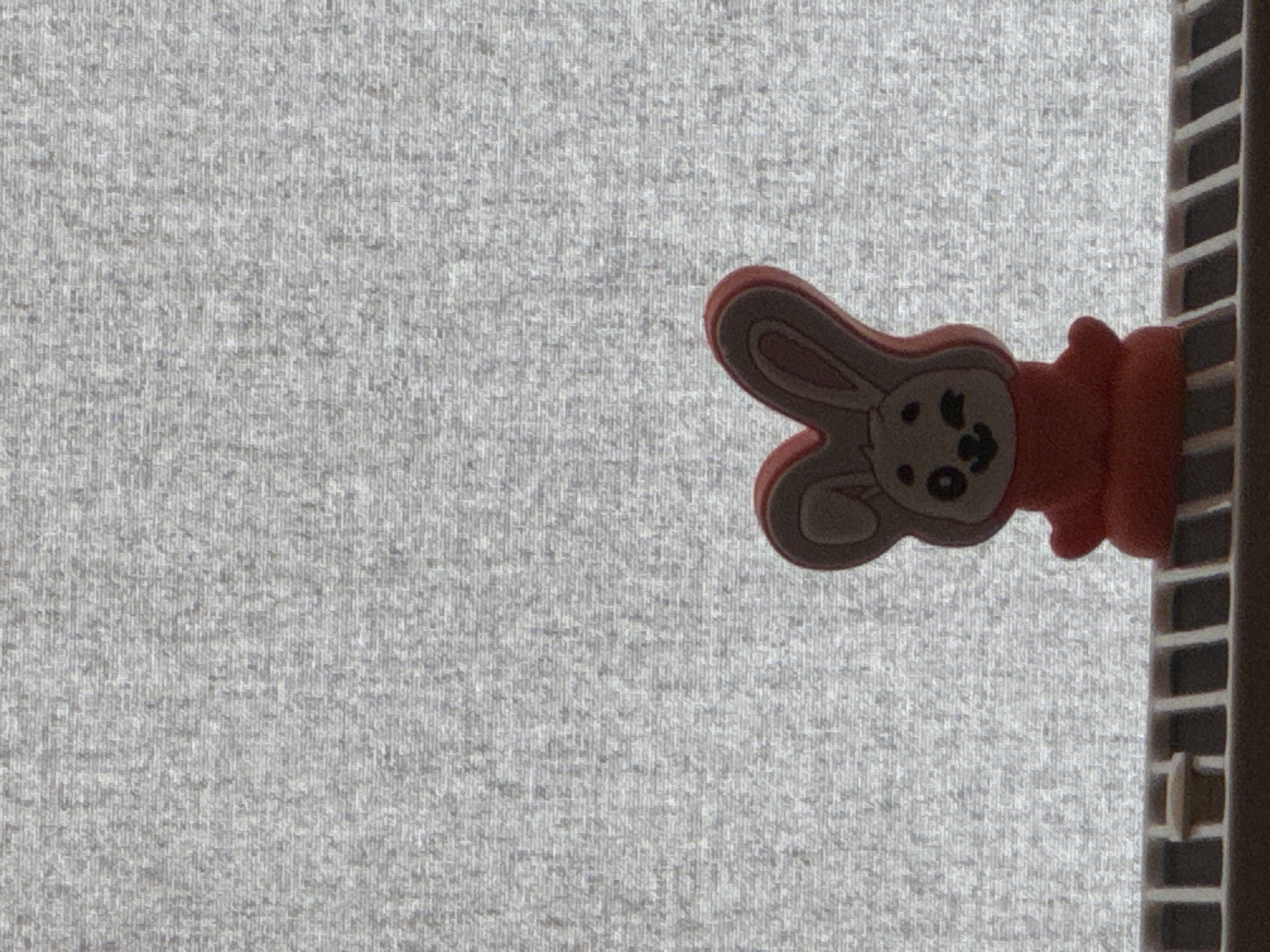 A small, red, cartoonish rabbit figurine with brown ears is perched on a white, grid-like shelf against a textured gray background. The rabbit is depicted from the waist up, with one eye closed in a wink. Its body is round, and its head features a simple smiley face. The background appears to be fabric, possibly a curtain or blinds, with a uniform, grainy texture. The lighting suggests the source is behind the background, creating a silhouette effect on the figurine and highlighting its form. The shelf is made of plastic or a similar material and has a repeating pattern of rectangular openings. The scene is static and appears to be an indoor setting. No city or specific location is identifiable from the image.