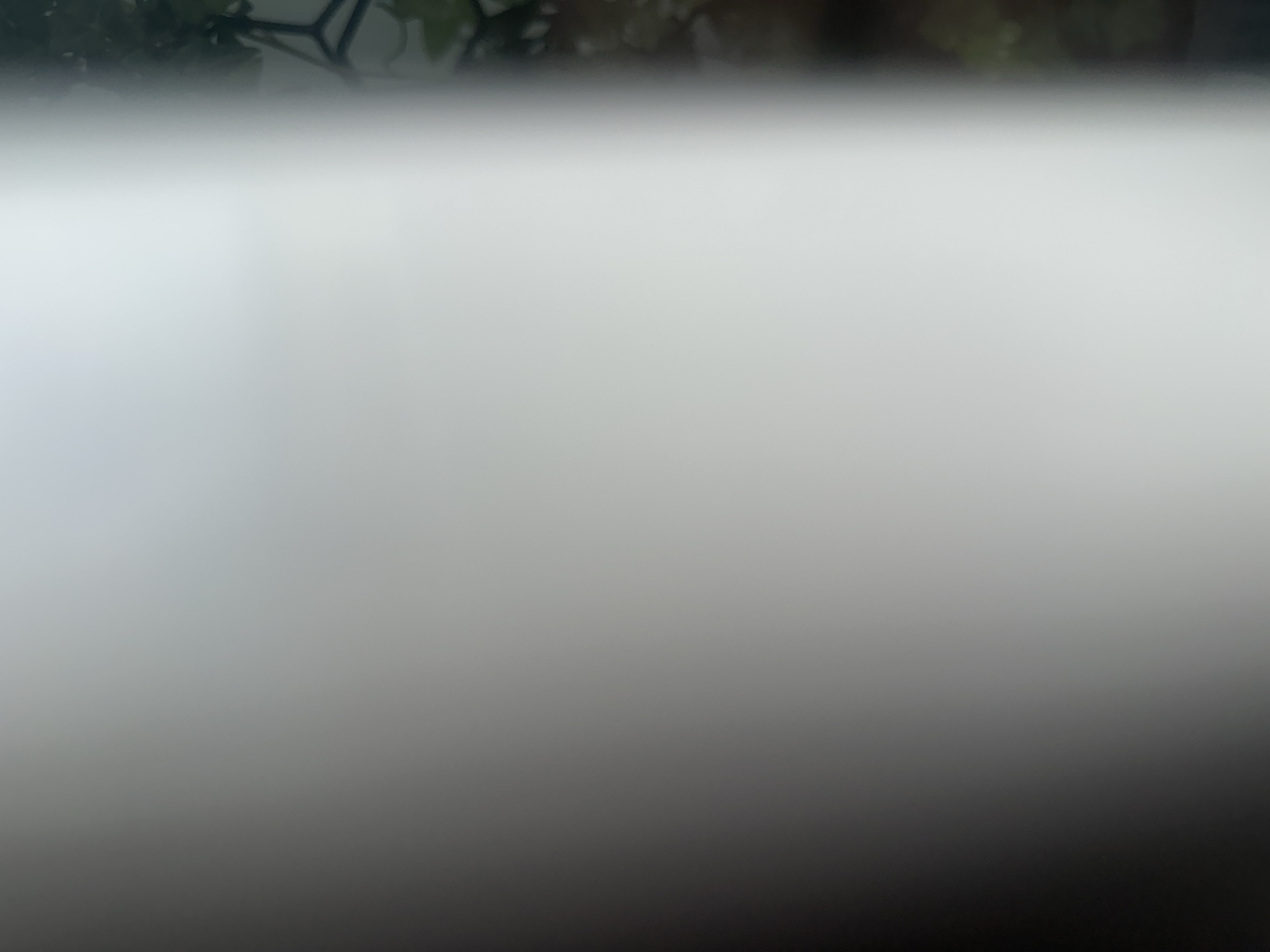The image is a blurry, out-of-focus photograph. A large, bright, out-of-focus white object dominates the left side of the frame, obscuring most of the scene. To the right of this white object, a dark, blurry background with hints of green foliage and possibly some metallic structures is visible. The scene's location in Belgrade, Serbia, cannot be determined from the provided image due to the extreme lack of detail and focus. There are no discernible people, subjects, actions, or interactions depicted.